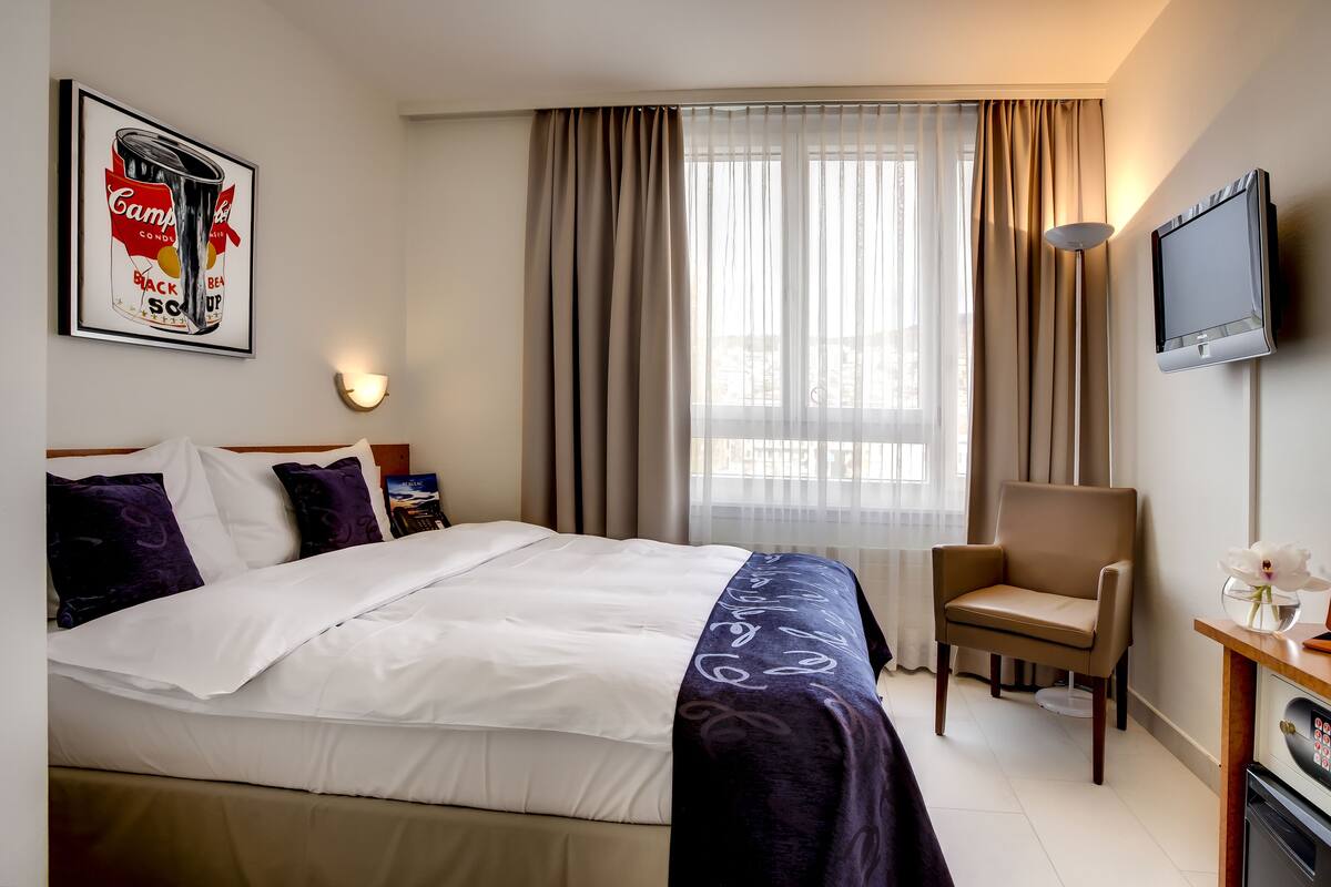 A hotel room with a bed, a chair, a desk with a television, and a window with curtains.