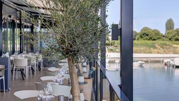 Un espacio al aire libre con mesas y sillas, un árbol y vistas a una masa de agua y edificios al fondo.