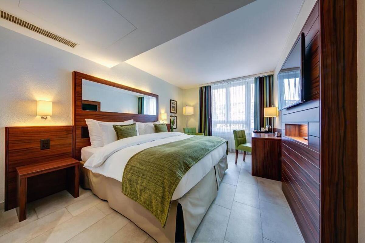 A hotel room with a large bed, bedside tables, a desk, a TV, and a window with curtains.