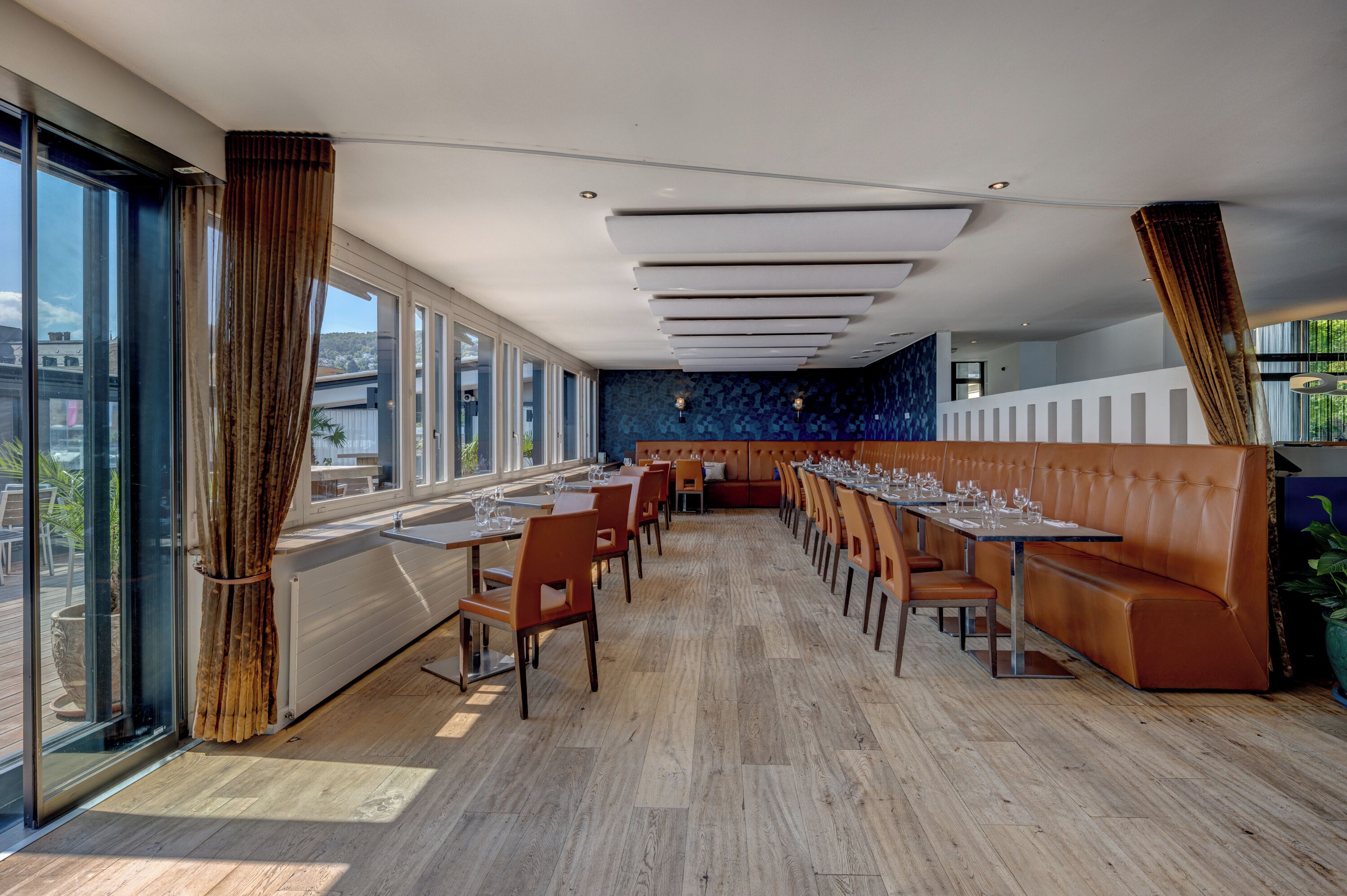 A modern restaurant with a long counter, wooden flooring, and large windows offering a city view.