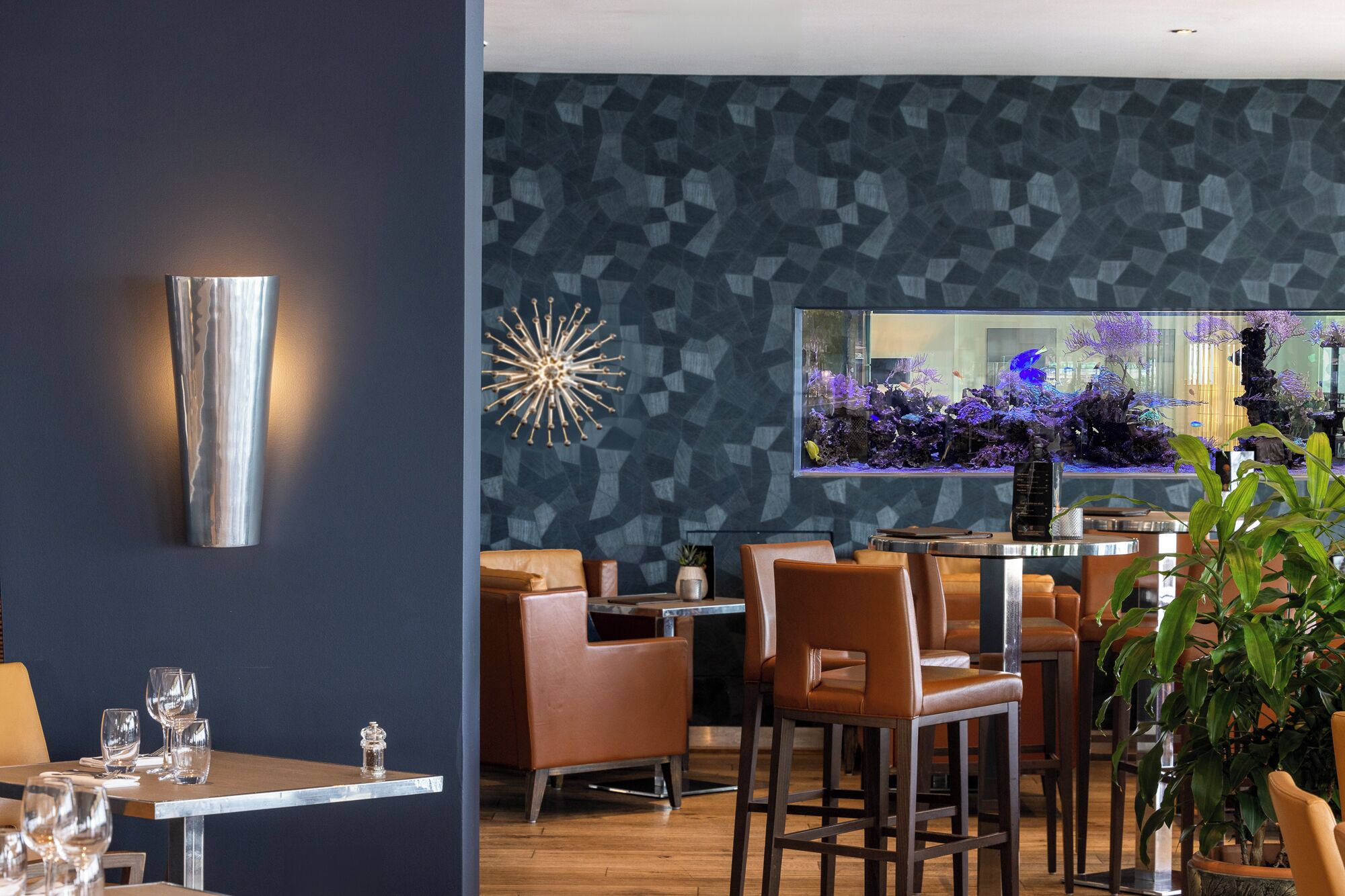 A modern bar area with leather seating, a decorative wall with a geometric pattern, and a large aquarium with colorful fish.