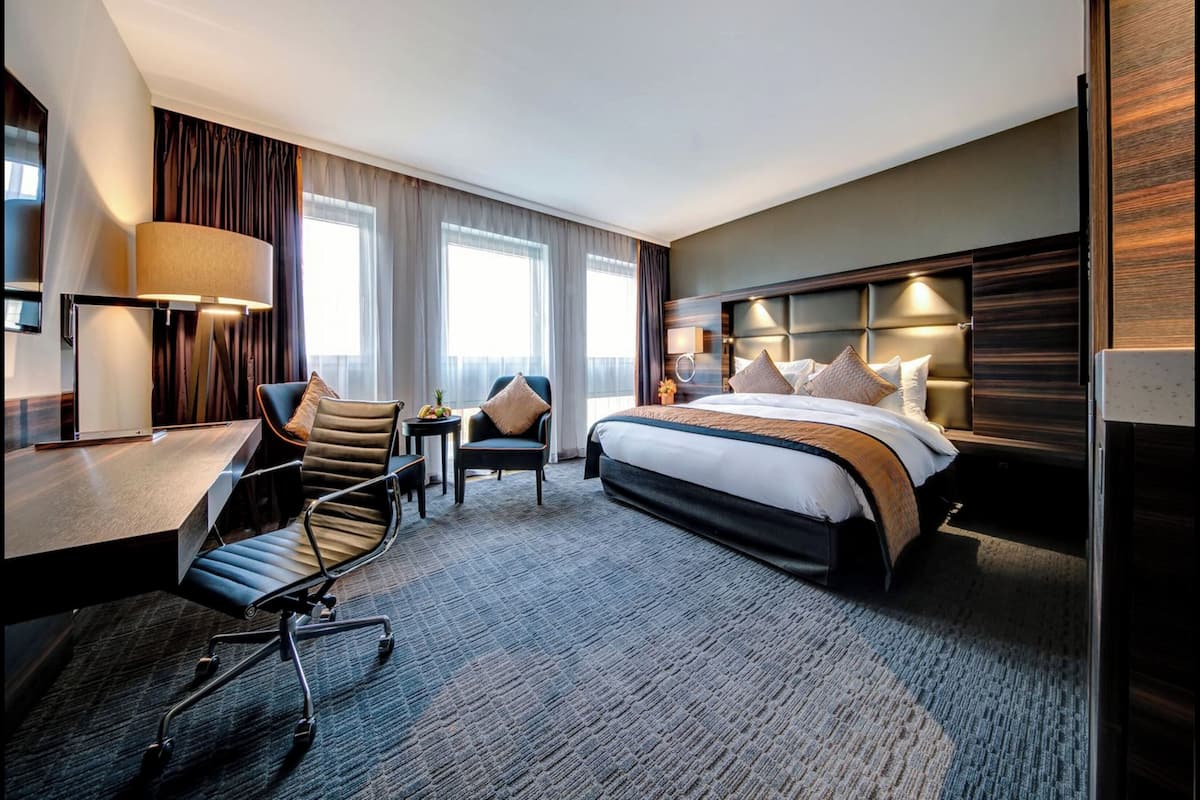 A hotel room with a bed, desk, chair, and a small table with a lamp.