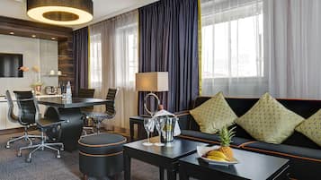 A modern hotel room with a dining area, a sofa with patterned cushions, a coffee table with fruit, and a large window with curtains.