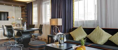 Una habitación de hotel moderna con zona de comedor, un sofá con cojines estampados, una mesa de centro con fruta y un amplio ventanal con cortinas.