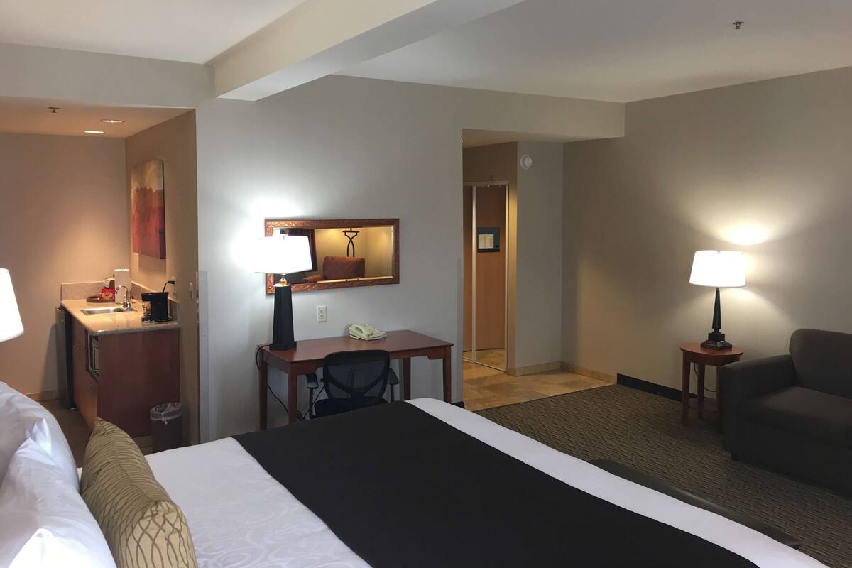 A hotel room with a bed, a desk with a chair, a small table with a lamp, a sofa, and a bathroom with a sink and mirror.