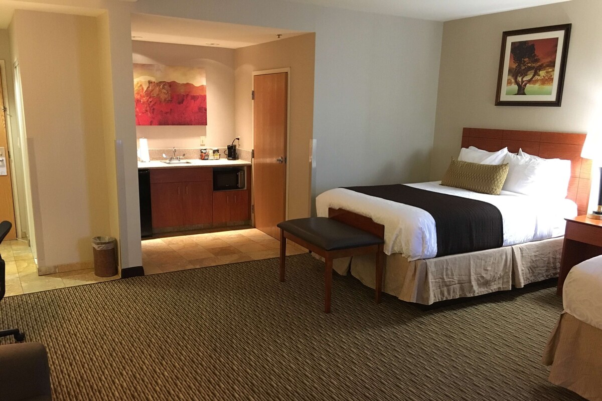 A hotel room with a bed, a bench, a kitchenette, and a painting on the wall.