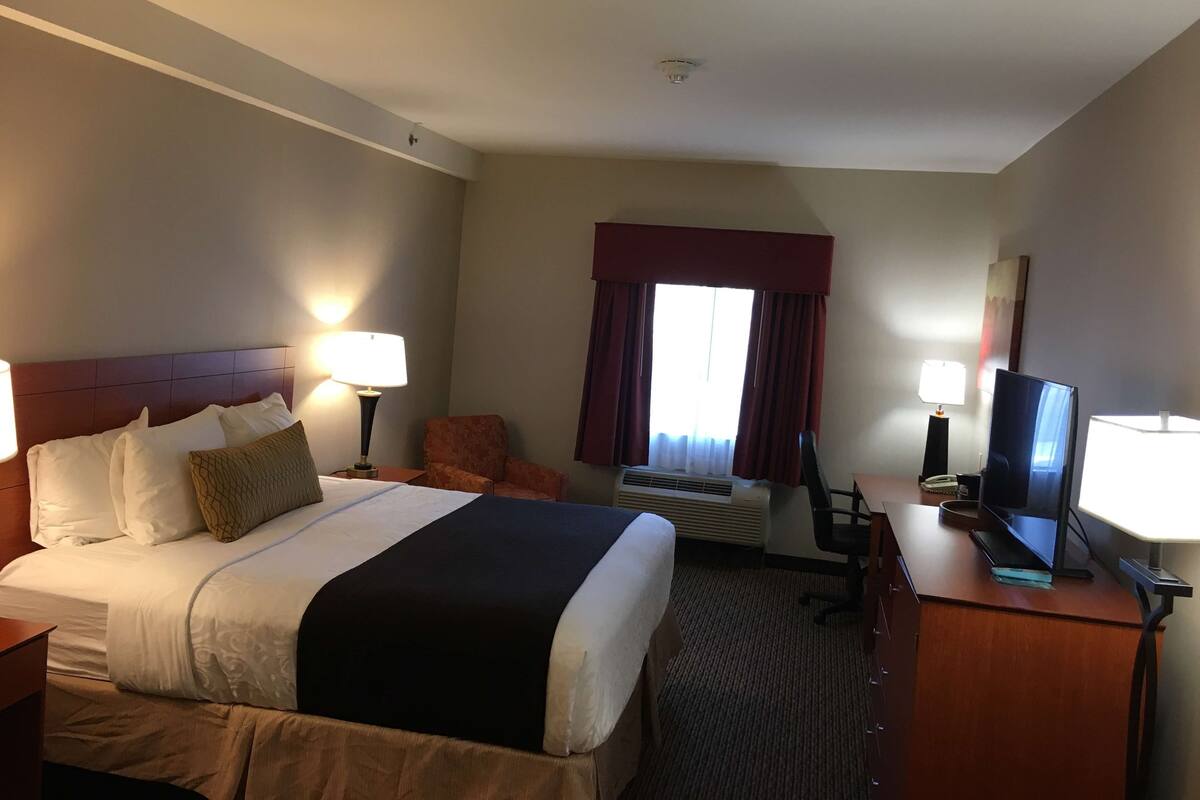 A hotel room with a bed, two bedside lamps, a chair, a desk with a television, and a window with curtains.