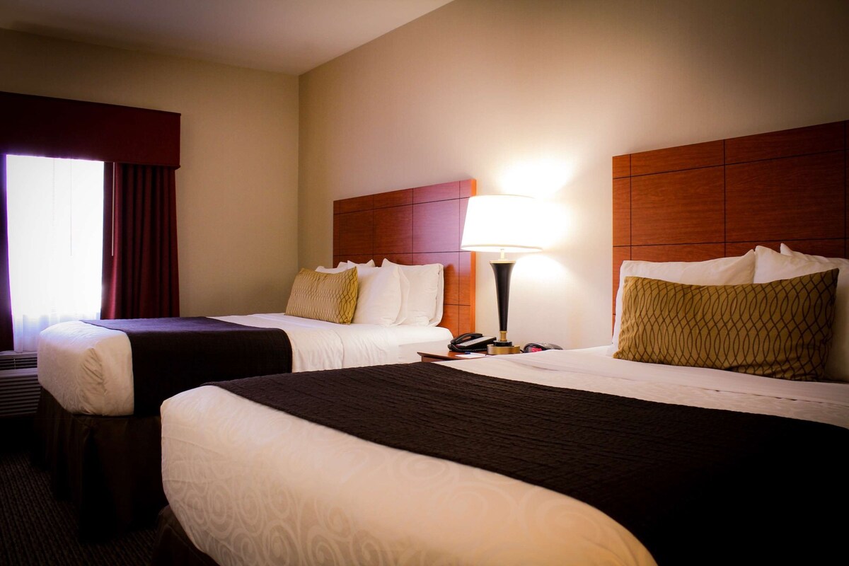 A hotel room with two beds, a nightstand with a lamp, and a window with curtains.