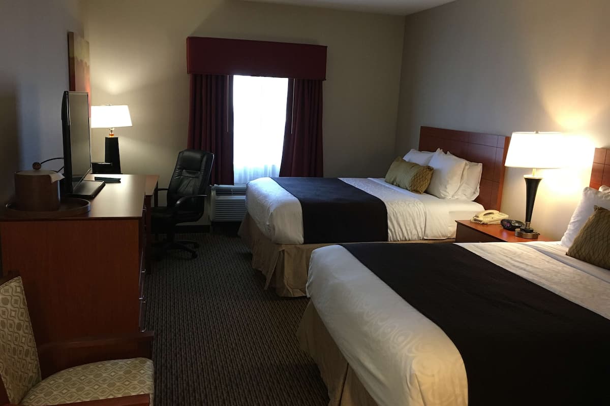 A hotel room with two beds, a desk with a lamp, a chair, a TV, and an air conditioning unit.