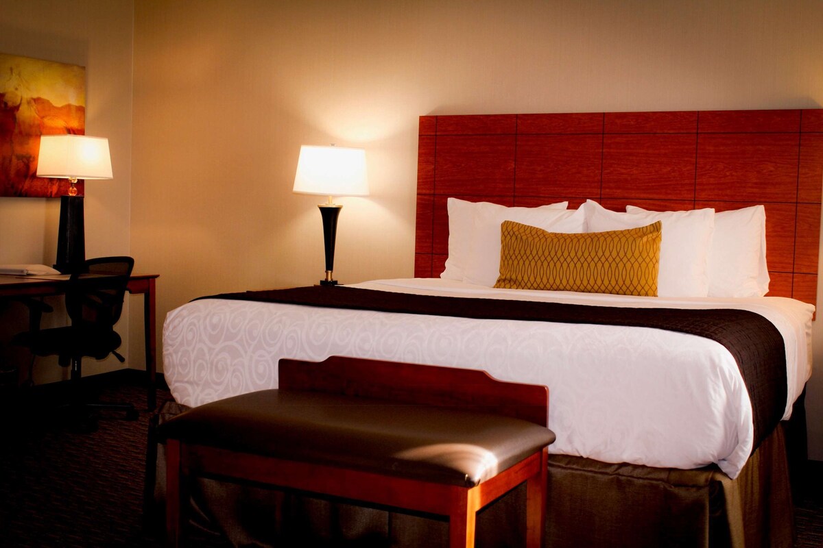 A hotel room with a large bed, a desk with a chair, a lamp, and a bench.