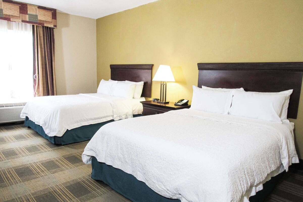 A hotel room with two beds, a nightstand with a lamp and phone, and a window with curtains.
