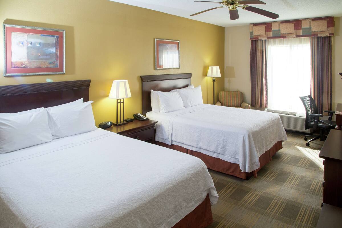 A hotel room with two beds, a ceiling fan, a desk, and a chair.
