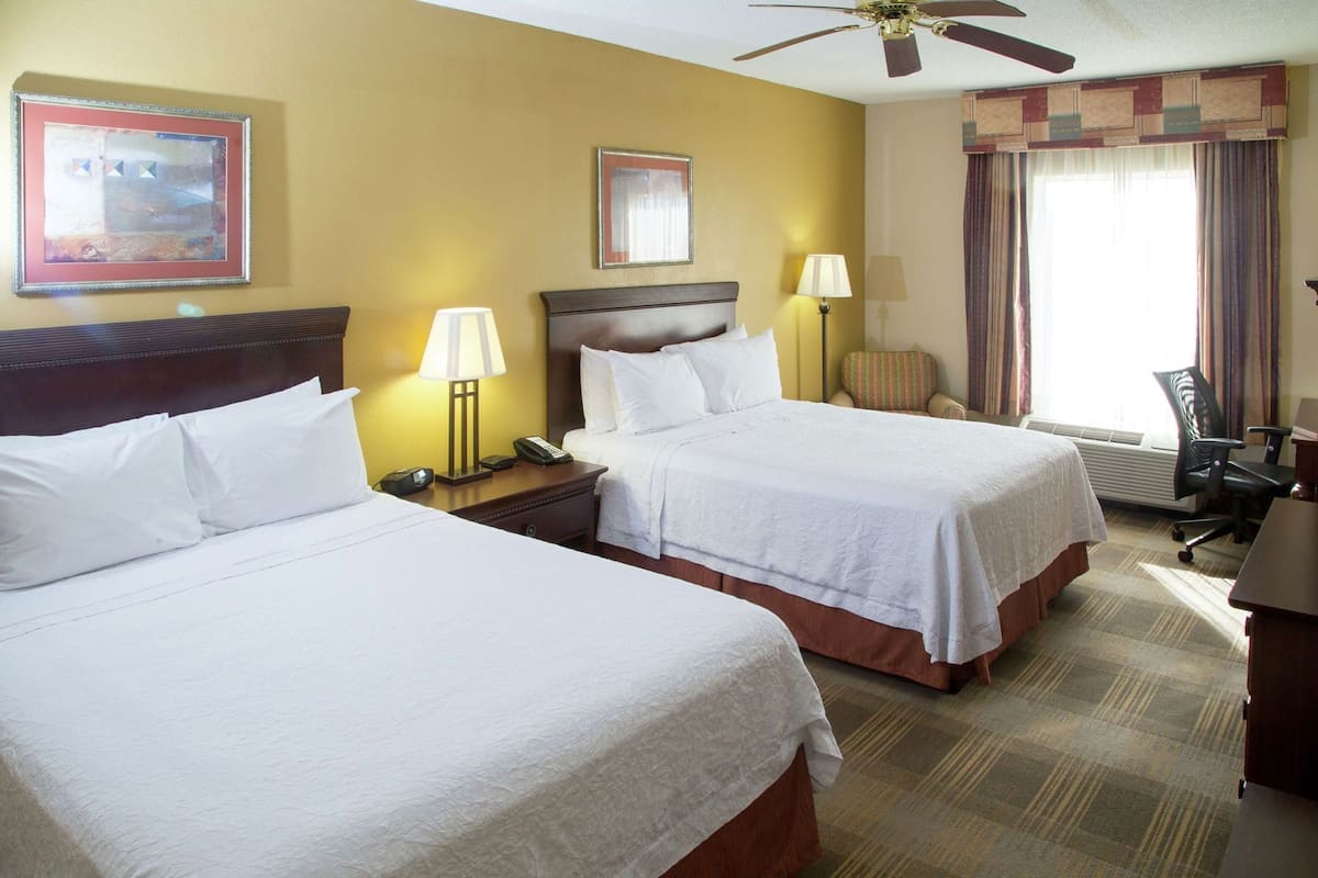A hotel room with two beds, a desk, a chair, a ceiling fan, and two lamps.