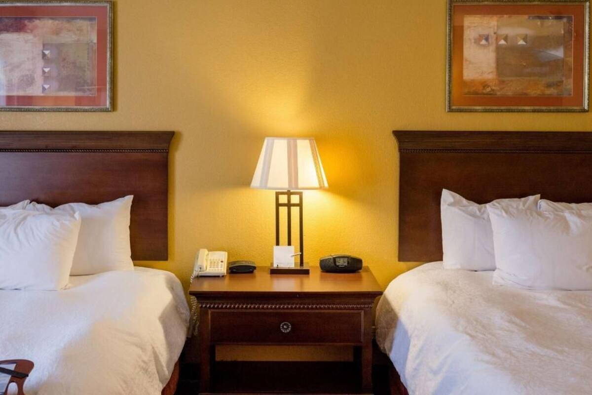 A hotel room with two beds, a nightstand with a lamp, a phone, and a small table.