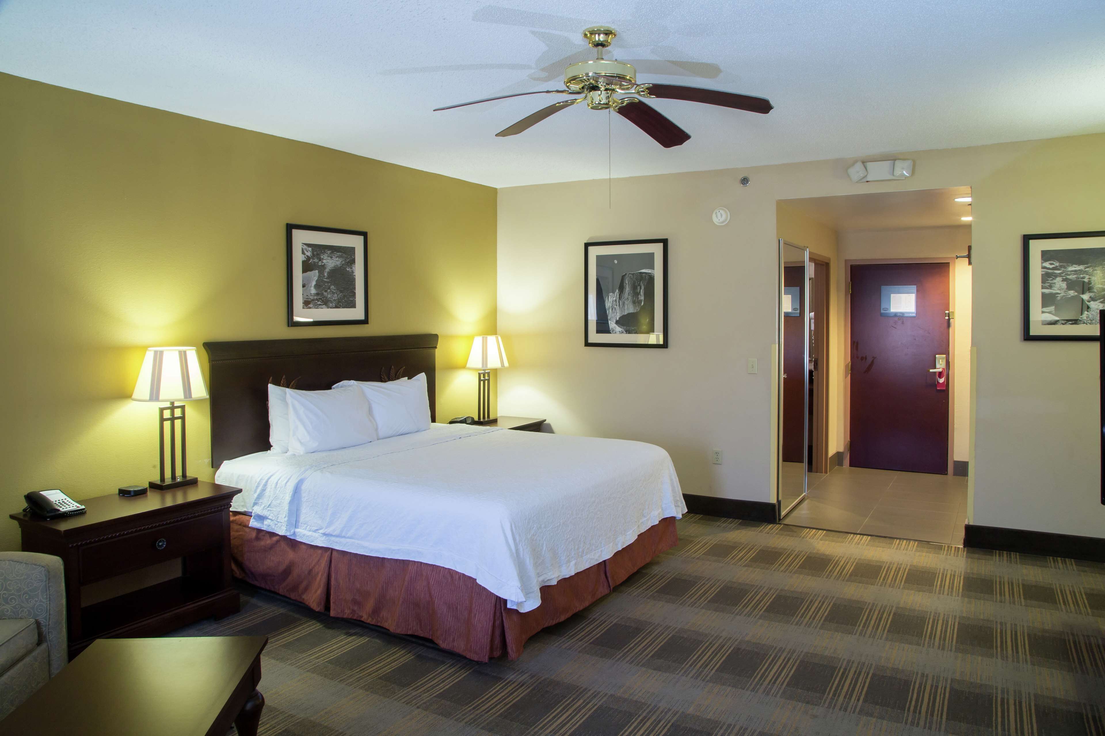 A hotel room with a bed, two bedside lamps, a nightstand, a ceiling fan, and framed pictures on the walls.