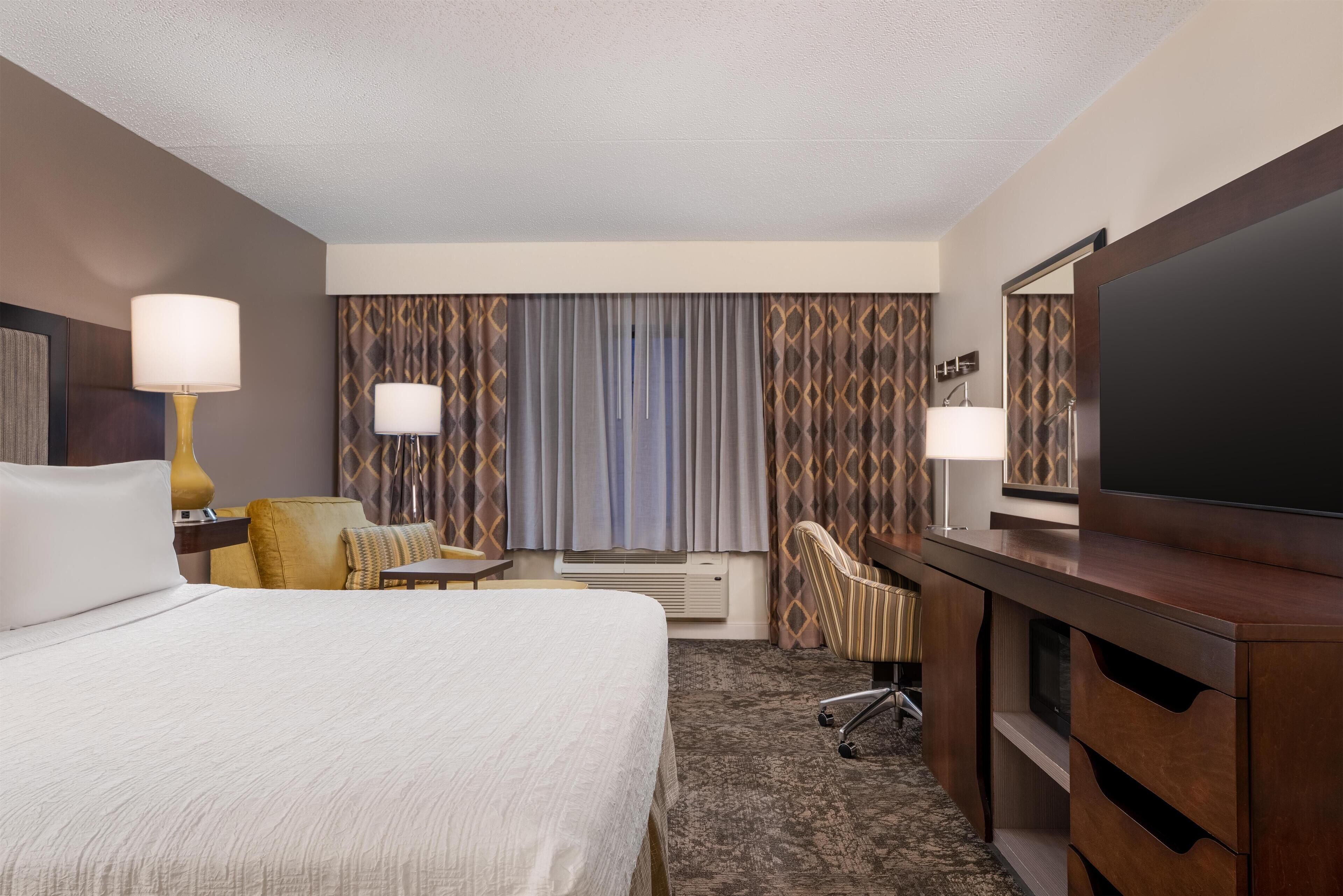 Quarto de hotel com uma cama grande, uma escrivaninha com abajur, uma televisão e uma cadeira.