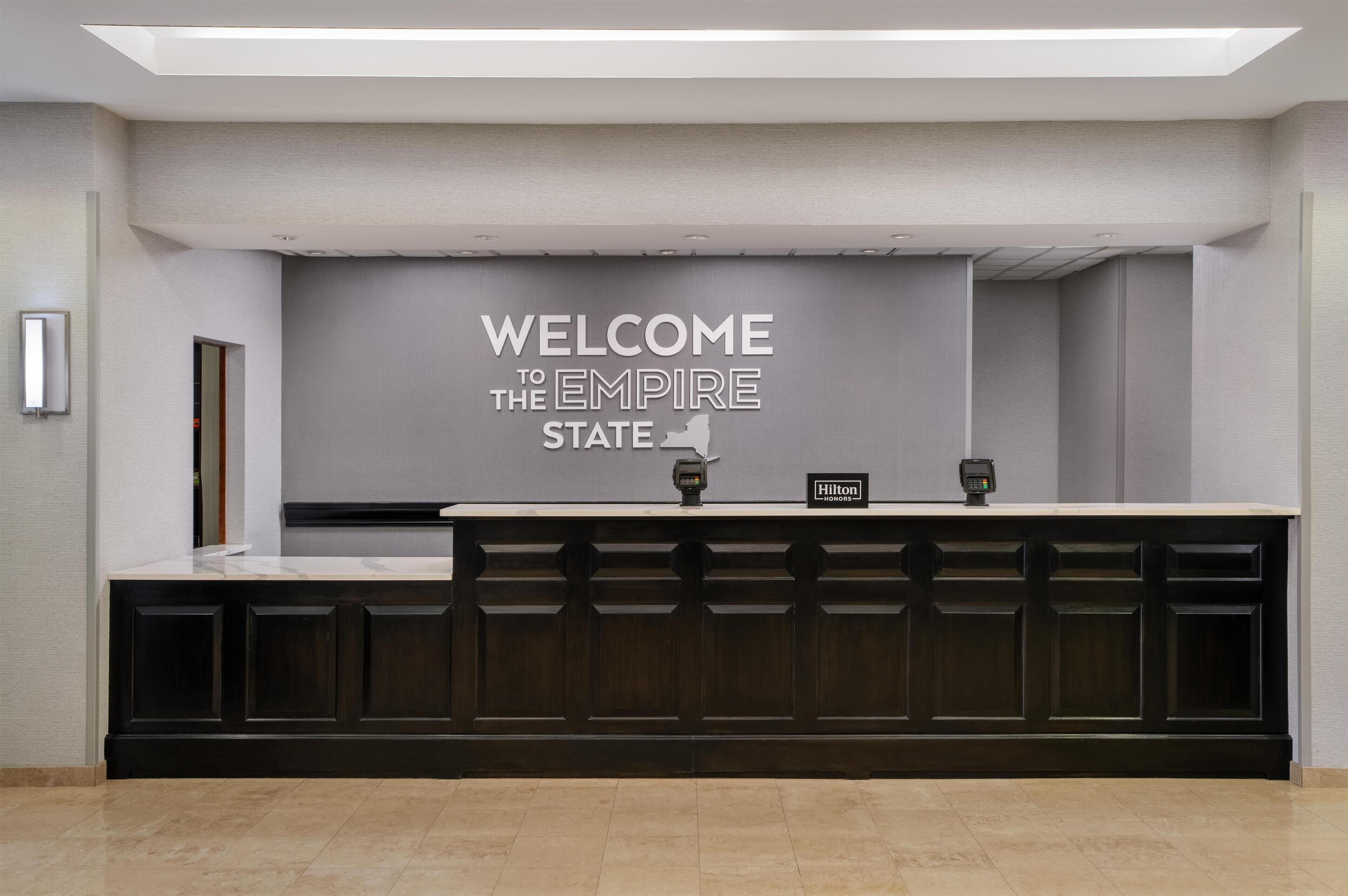 A reception desk with a "WELCOME TO THE EMPIRE STATE" sign, a "Hilton" sign, and a security camera.