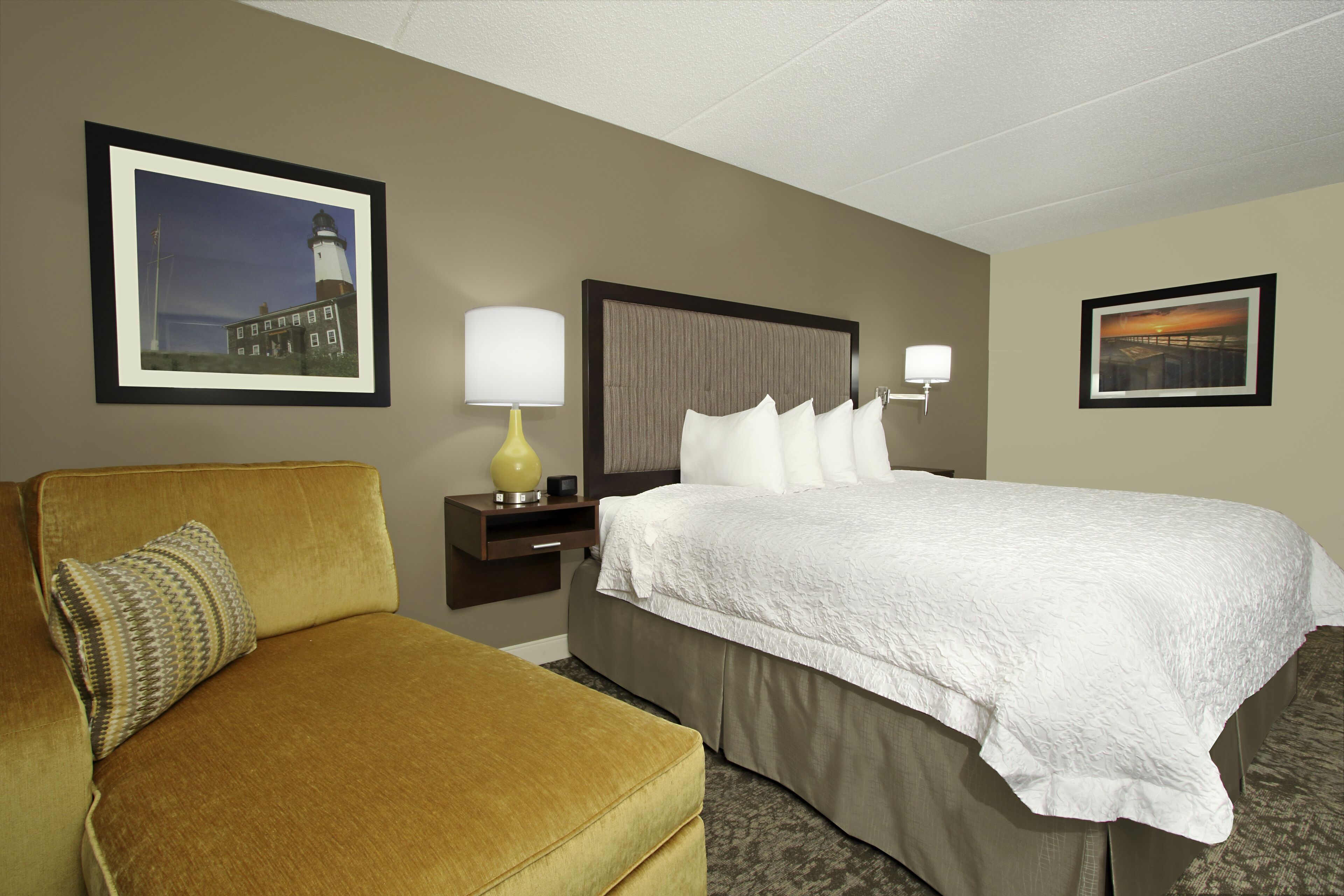 A hotel room with a bed, a sofa, a nightstand, a lamp, and two framed pictures on the walls.