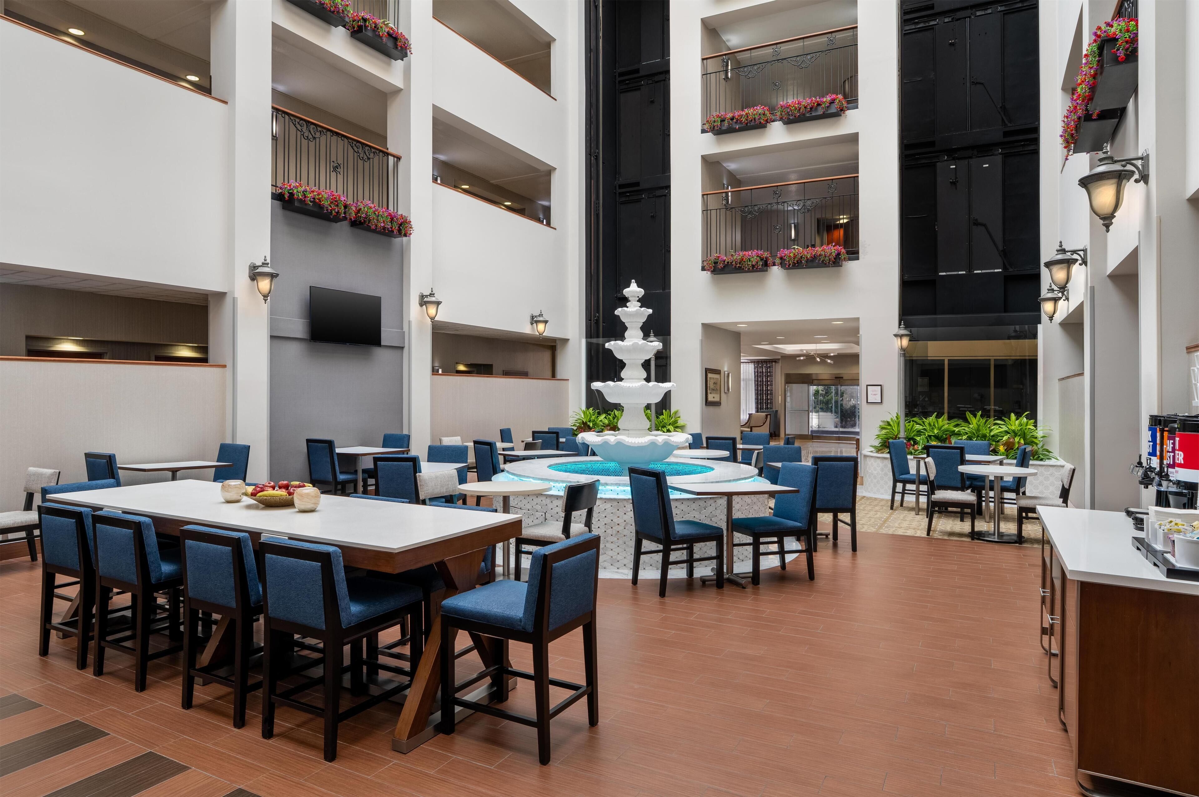 A spacious indoor dining area with multiple tables and chairs, a central fountain, and balconies with flower boxes.