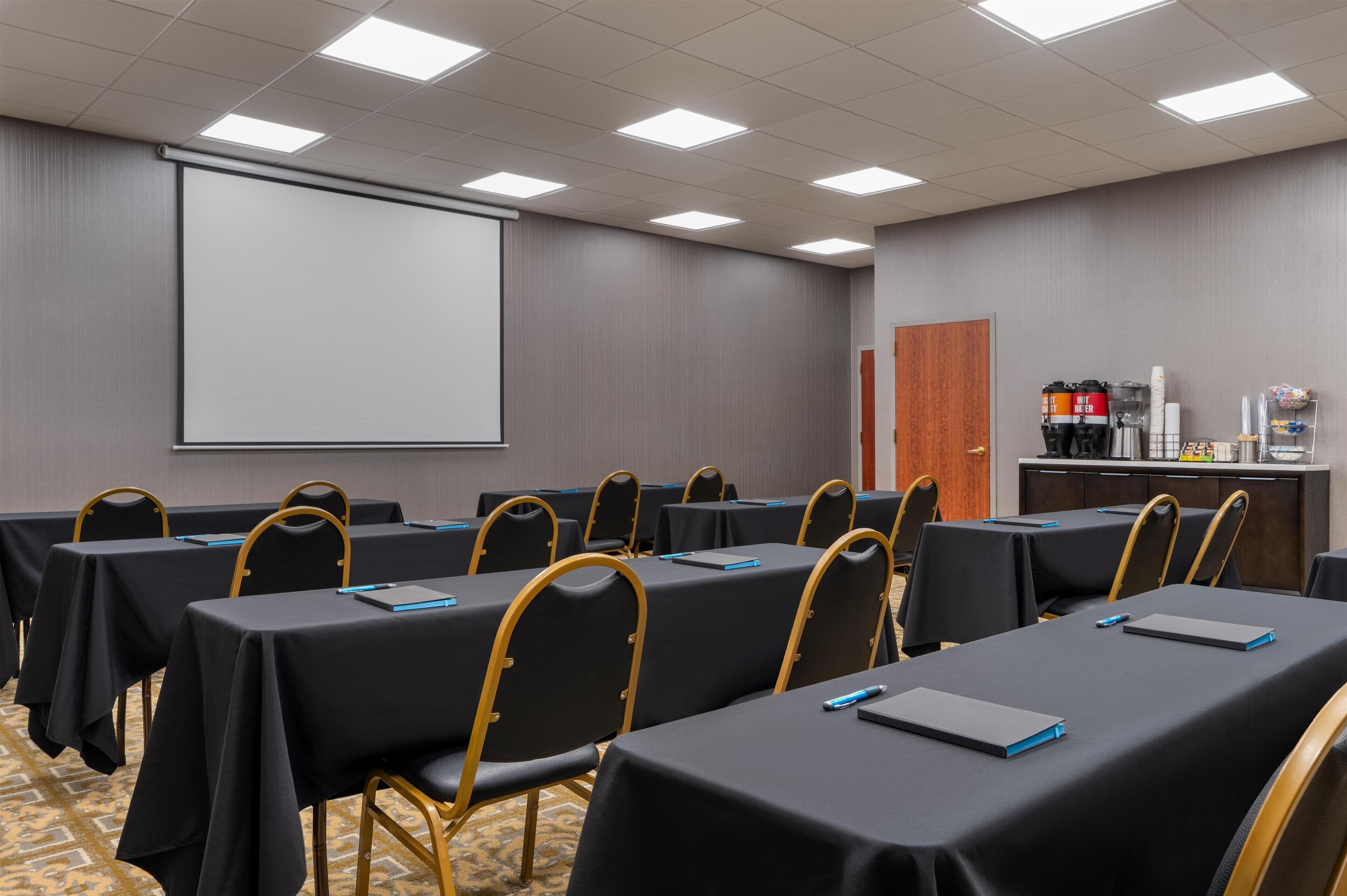 Ein Konferenzraum mit Tischen und Stühlen, einer Projektionsleinwand und einer Kaffeestation.