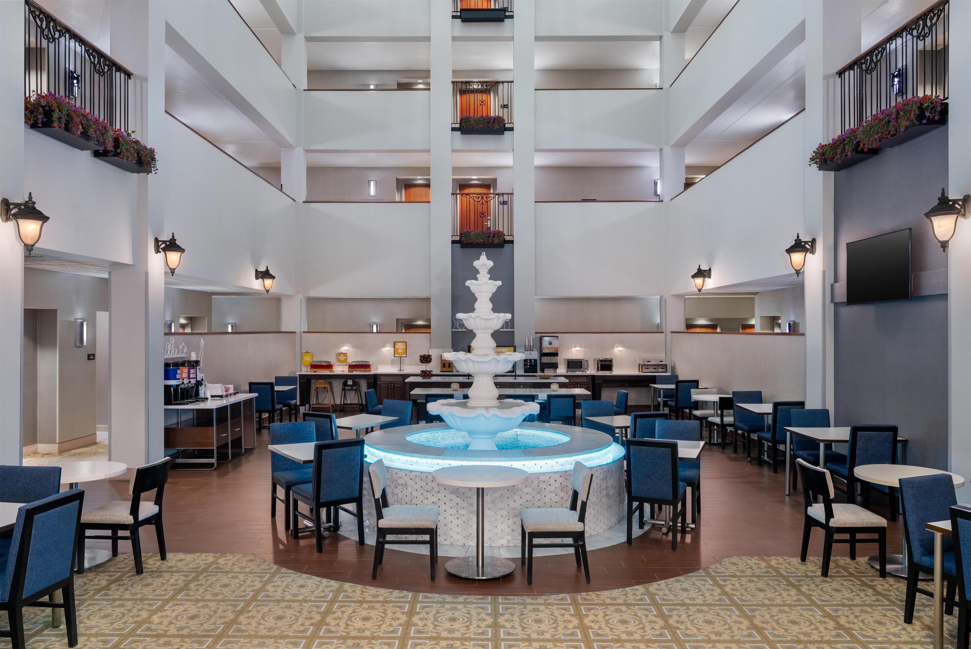 A spacious hotel lobby with a central fountain, multiple dining areas, and a modern staircase.