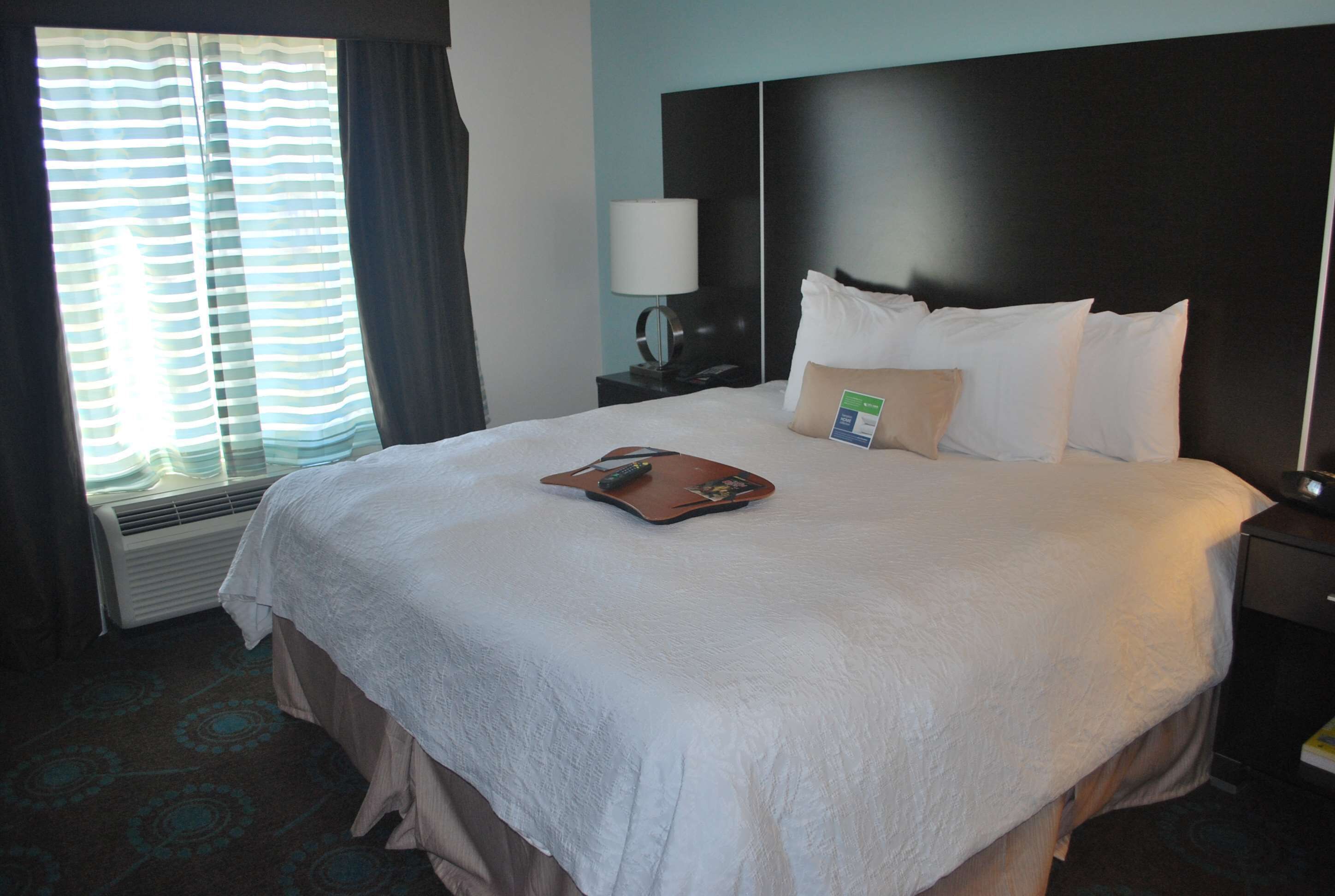 A neatly made bed with a pillow and a remote control on it, a bedside table with a lamp, and a window with curtains.