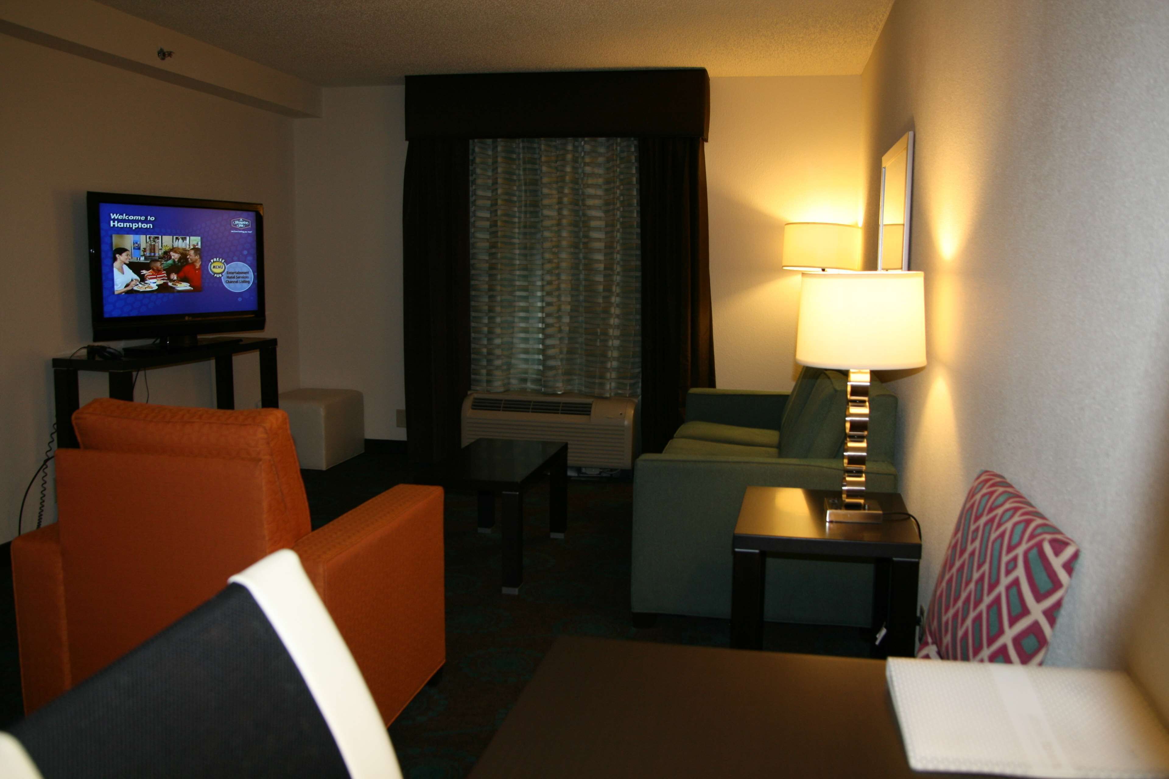 Ein Hotelzimmer mit Fernseher, der einen Begrüßungsbildschirm anzeigt, einer Couch, einem kleinen Tisch mit Lampe und einem Fenster mit Vorhängen.