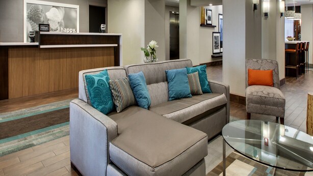A modern hotel lobby with a reception desk, a sectional sofa, a glass coffee table, and a dining area. - Hampton Inn Shreveport Airport (Shreveport)