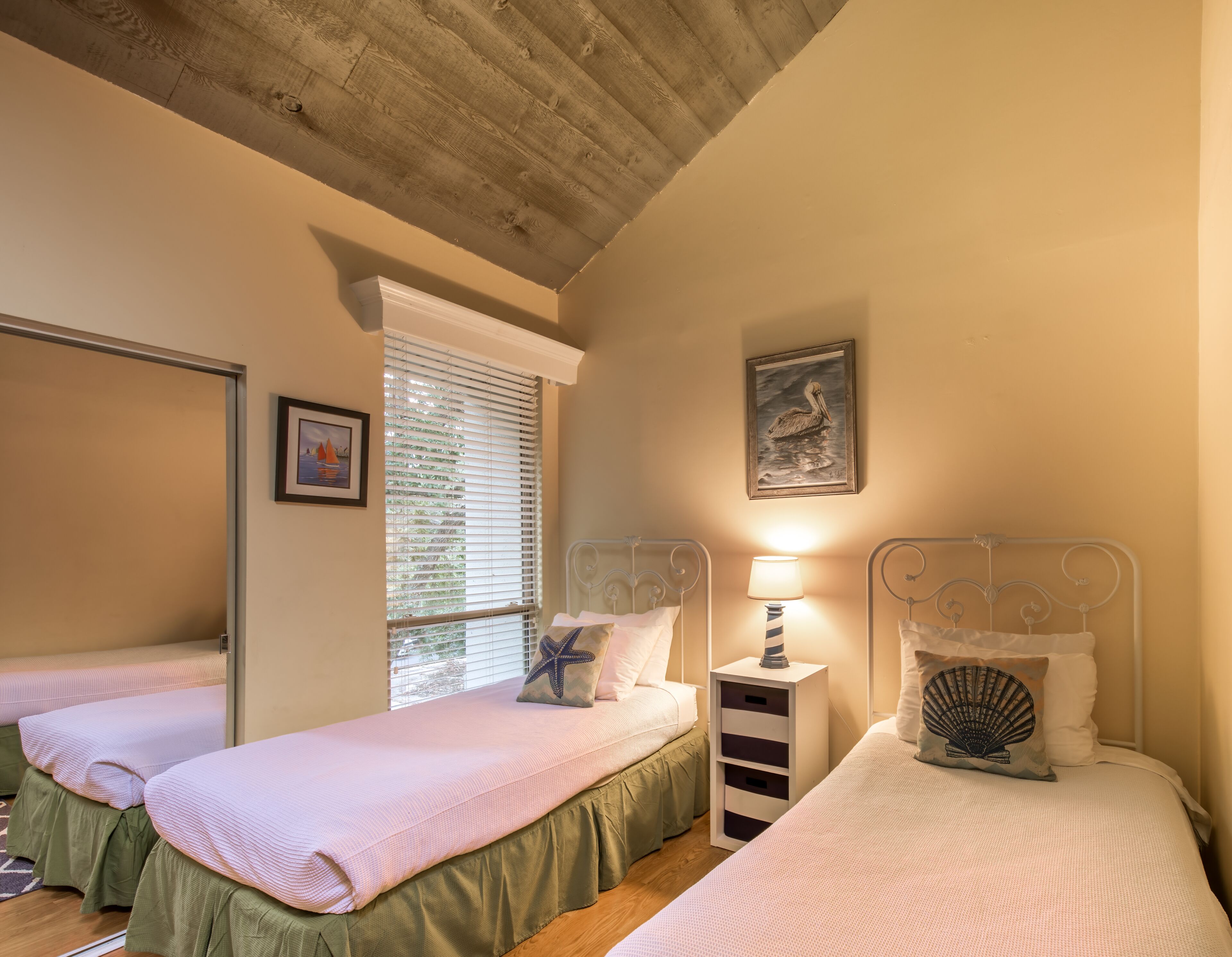 Une chambre avec deux lits simples, une table de chevet avec une lampe, un tableau encadré au mur et une fenêtre avec des stores.