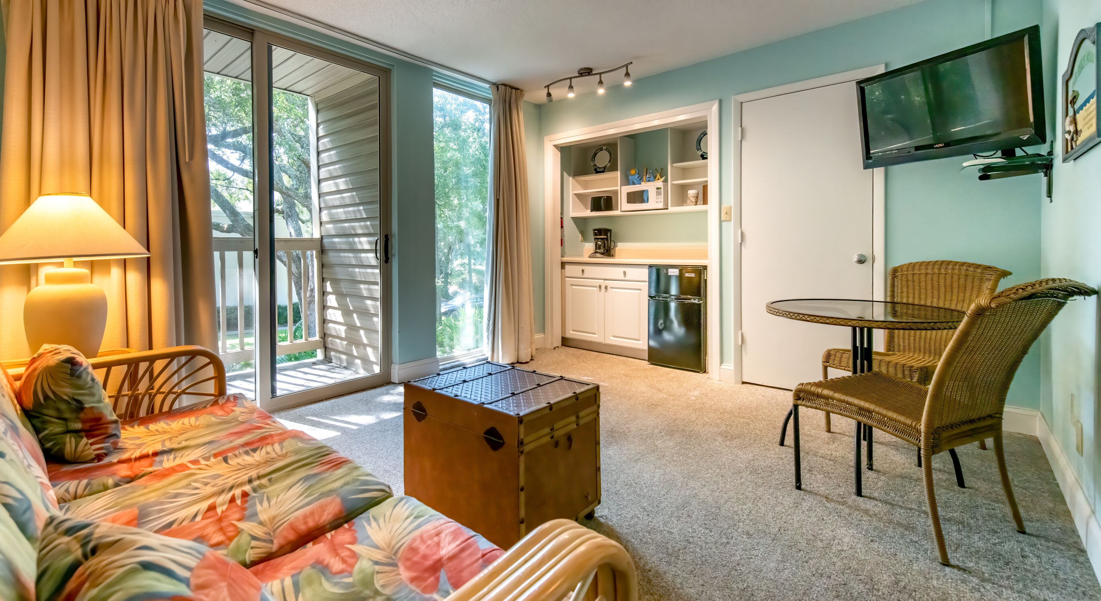 Una habitación acogedora con sofá, una mesita, una silla de mimbre, una zona de cocina con refrigerador y un televisor montado en la pared.
