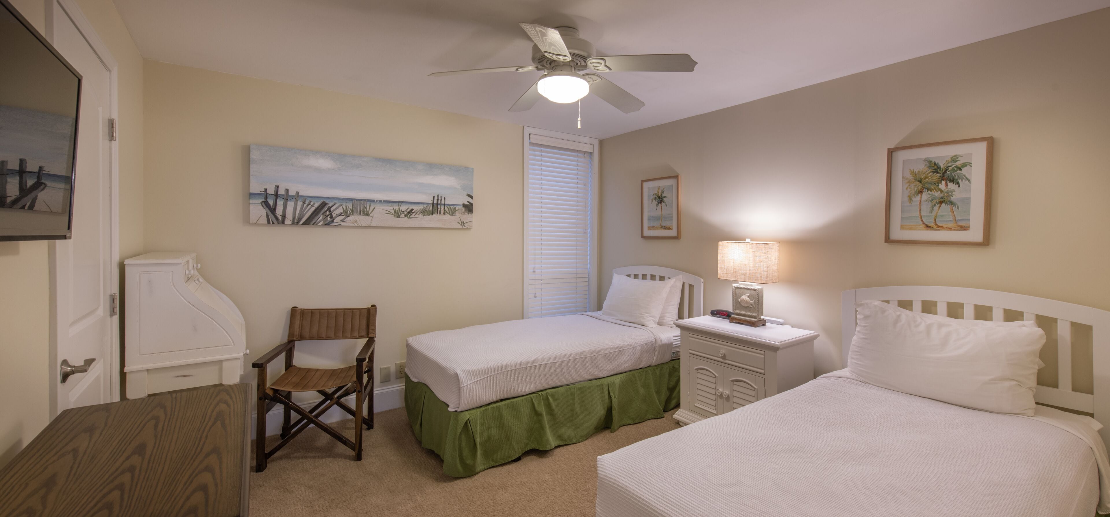 Une chambre avec deux lits simples, une chaise, une table de chevet avec une lampe, un ventilateur de plafond et des œuvres d’art accrochées aux murs.