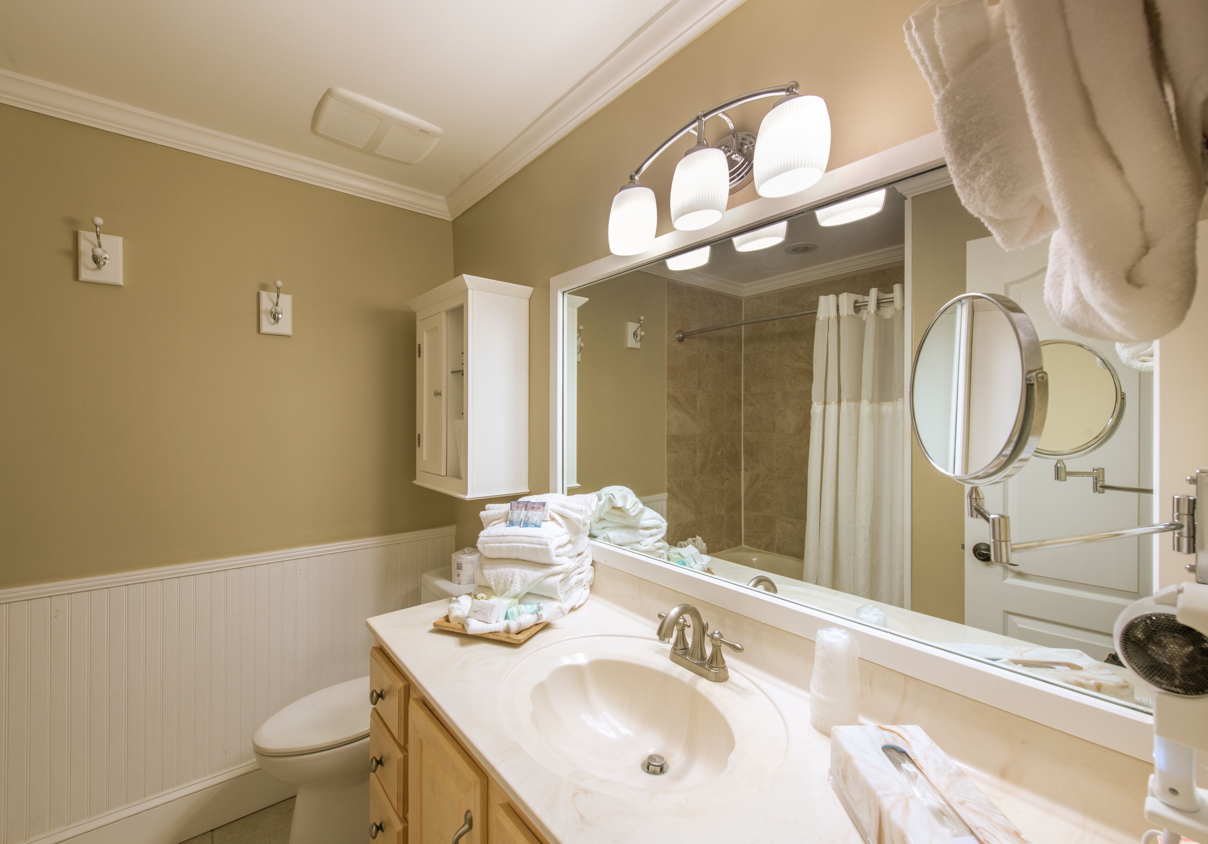 A bathroom with a sink, toilet, and a large mirror. There are towels stacked on the counter and a shower curtain visible in the background.