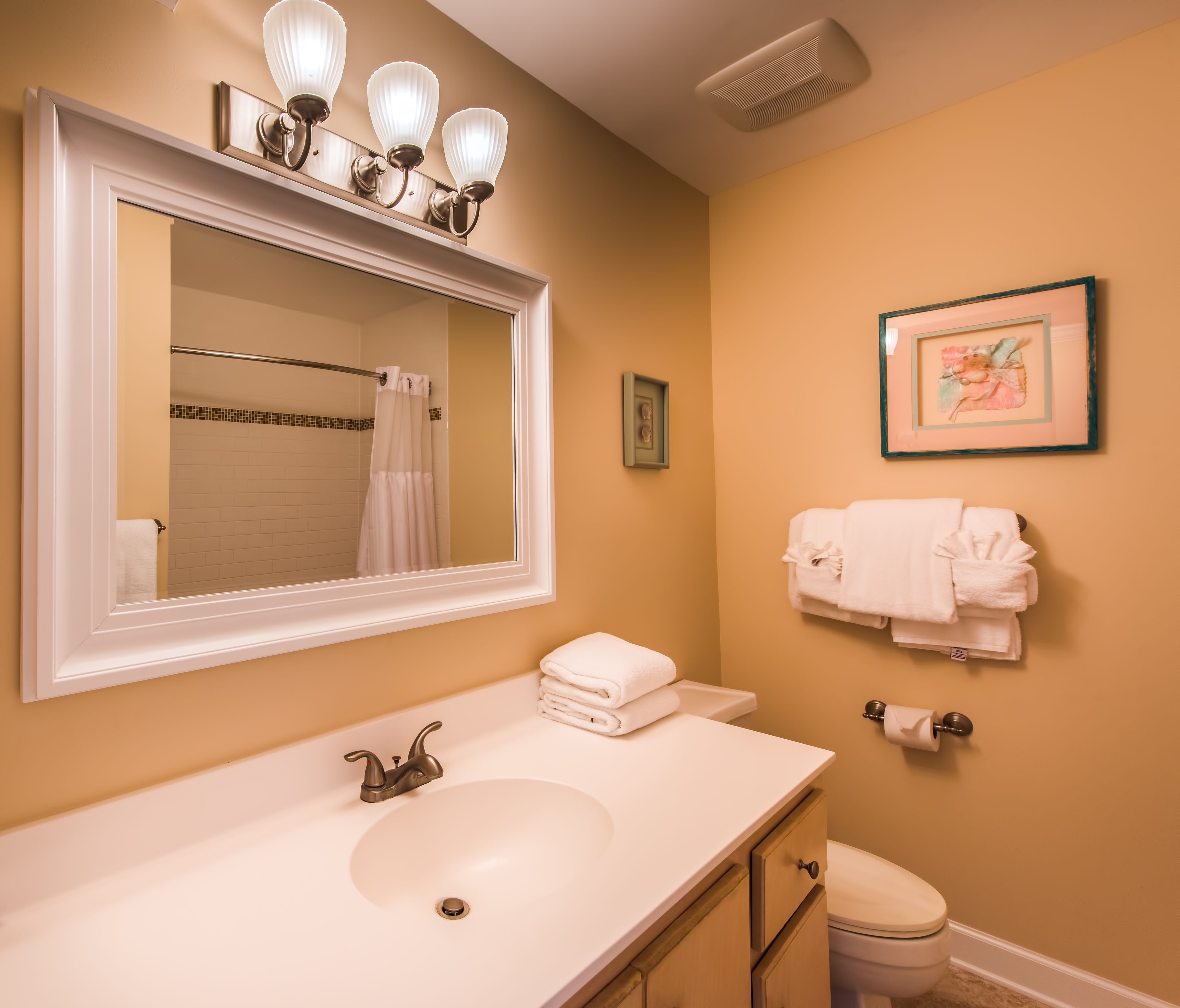 A bathroom with a white sink, a mirror with three lights, a toilet, and a framed picture on the wall.