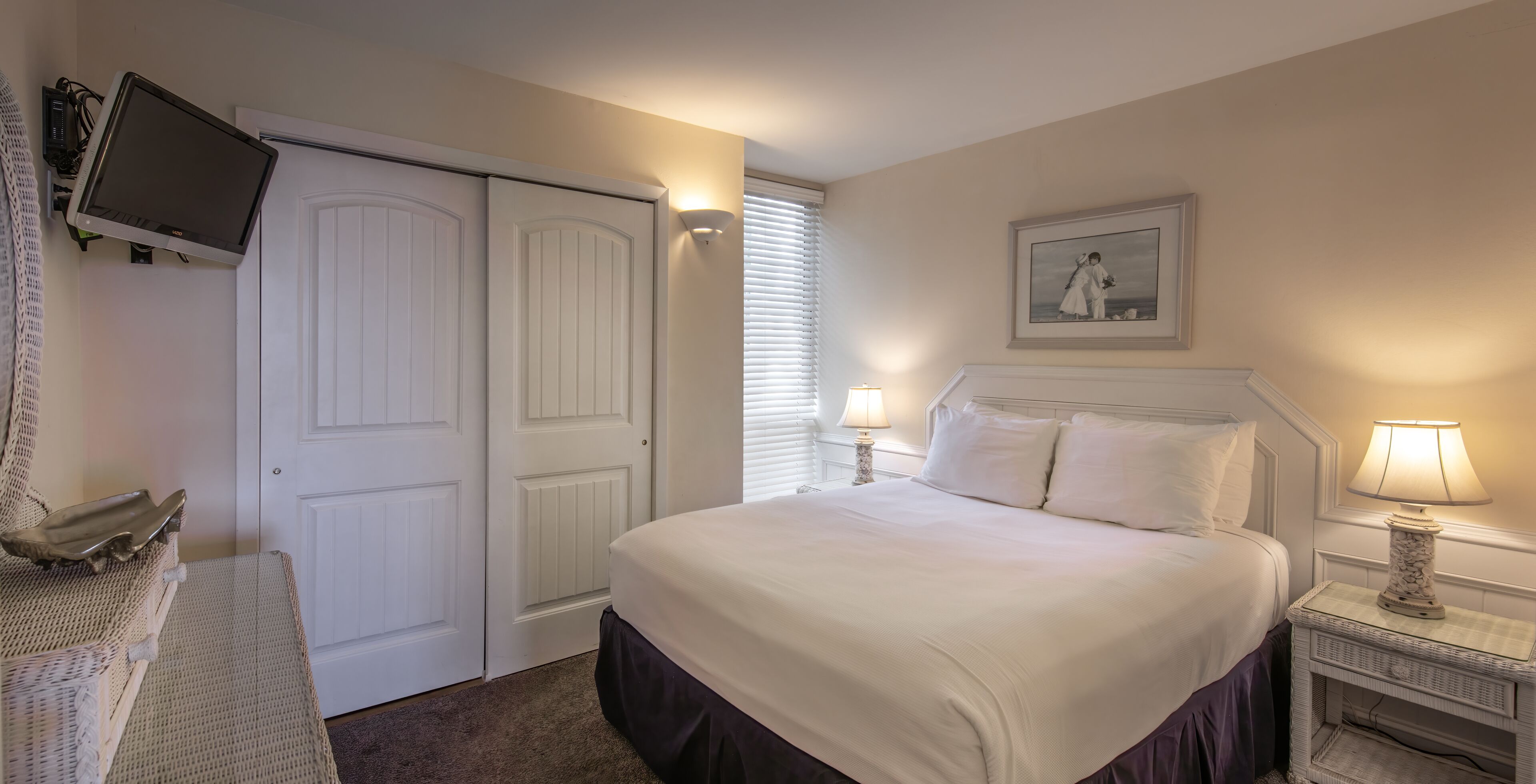 A neatly arranged bedroom with a bed, bedside lamps, a television mounted on the wall, and a framed picture on the wall.