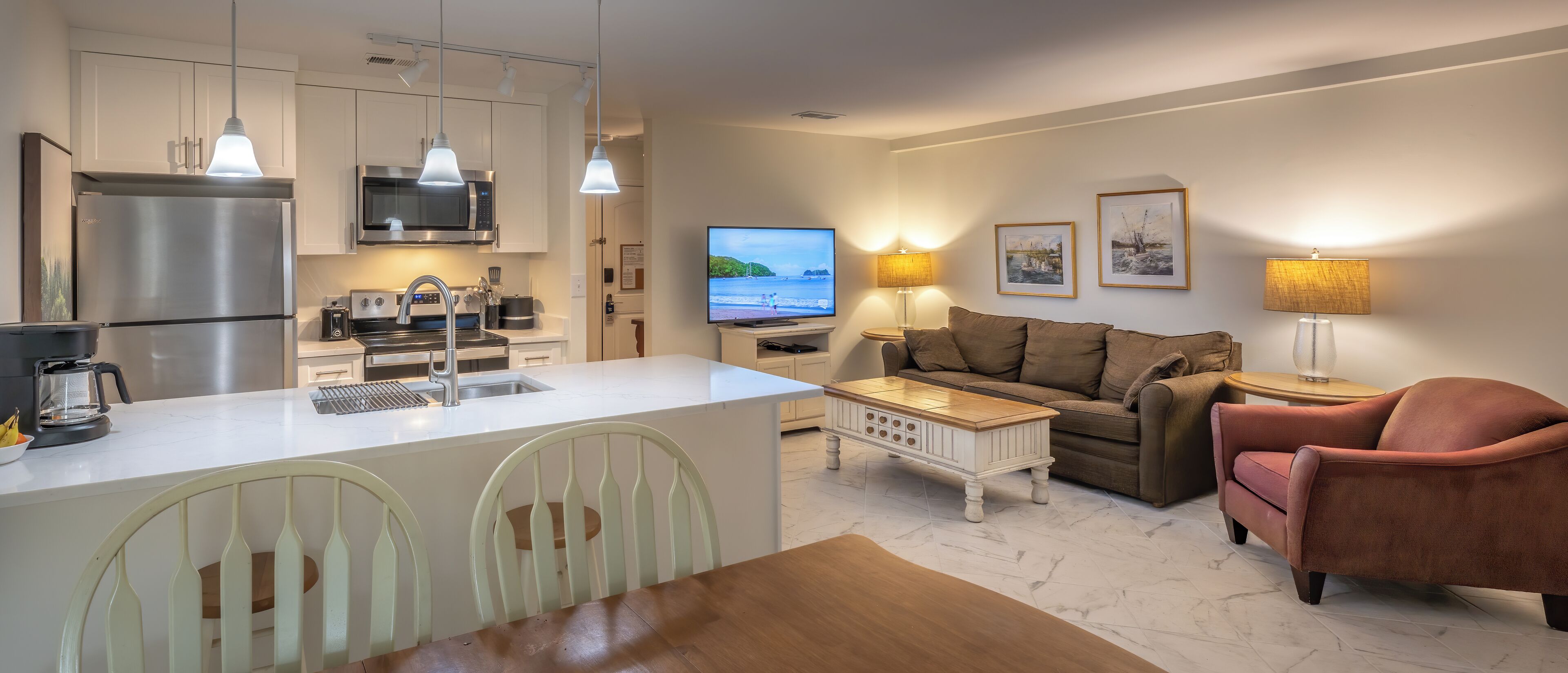 Una cocina moderna con refrigerador, microondas y cafetera. Hay un comedor con mesa y sillas, y una sala de estar con sofá y televisor.