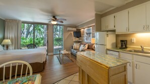 A compact hotel room with a bed, a small sofa, a kitchenette, and a view of greenery through the window. - Villas By the Sea Resort & Conference Center (Jekyll Island)