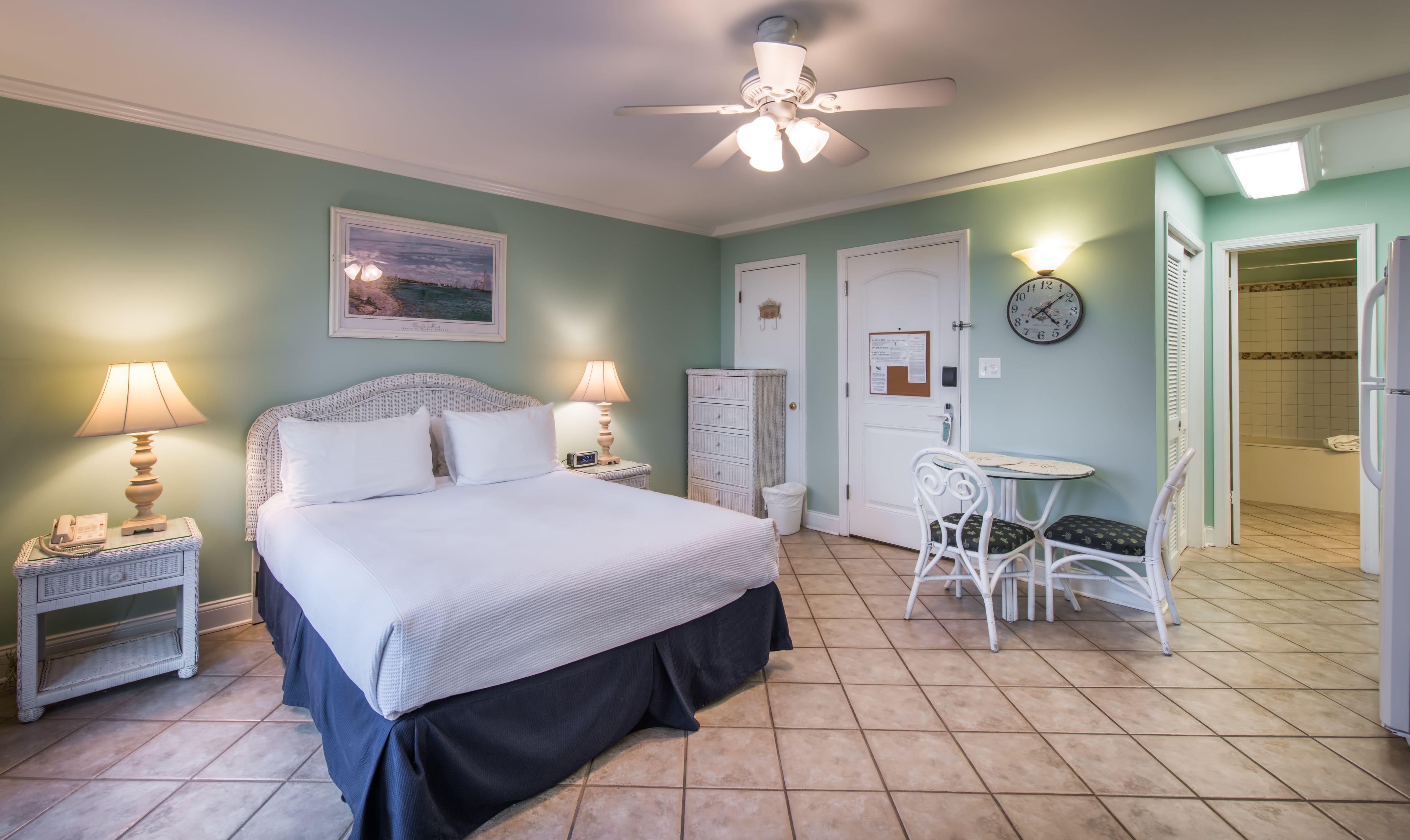 A bedroom with a bed, two bedside tables with lamps, a white chair, a small table, a ceiling fan, a clock, and a door to a bathroom.