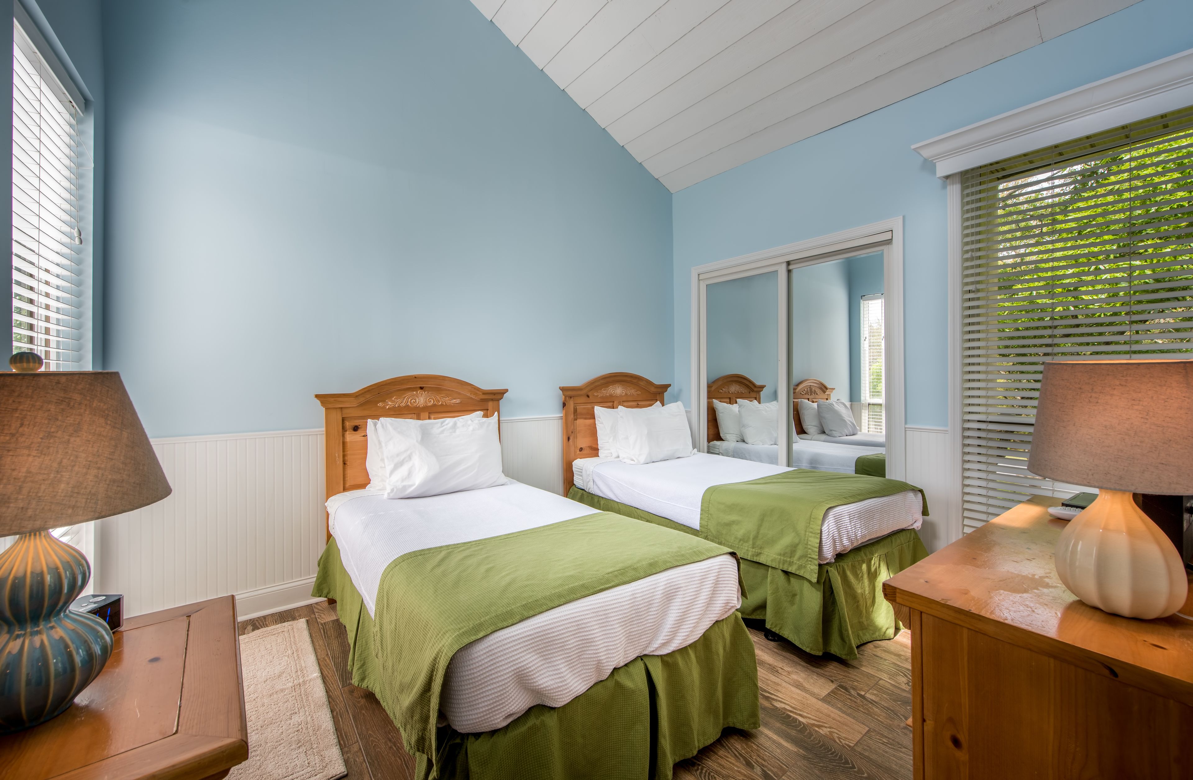 A bedroom with two beds, wooden headboards, white bedding, green bed skirts, a wooden nightstand with a lamp, a mirror, and a window with blinds.