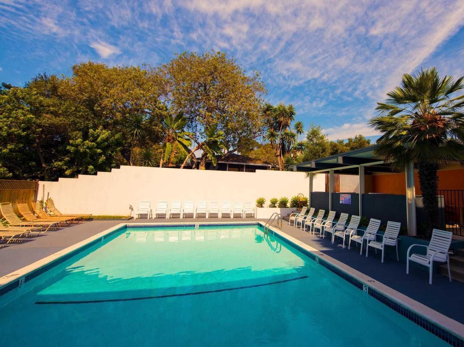 Une piscine rectangulaire à l’eau d’un bleu cristallin, entourée de chaises longues blanches et bordée d’un mur blanc.