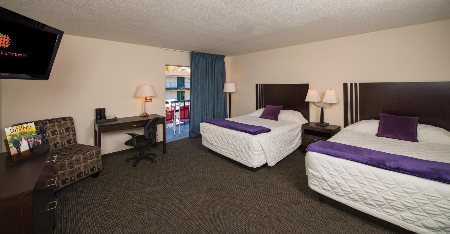A hotel room with two beds, a desk, a chair, a TV, and a dining area.