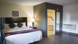Une chambre d’hôtel avec un grand lit, une table de chevet, une lampe, un tableau au mur et une salle de bain visible à travers une porte ouverte.