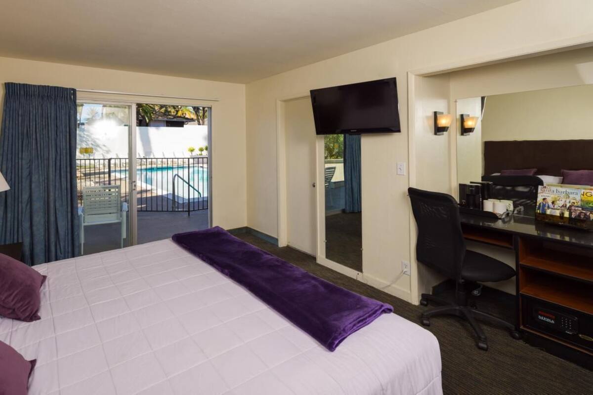 A hotel room with a bed, a desk, a chair, a TV, and a sliding door to a balcony with a pool.