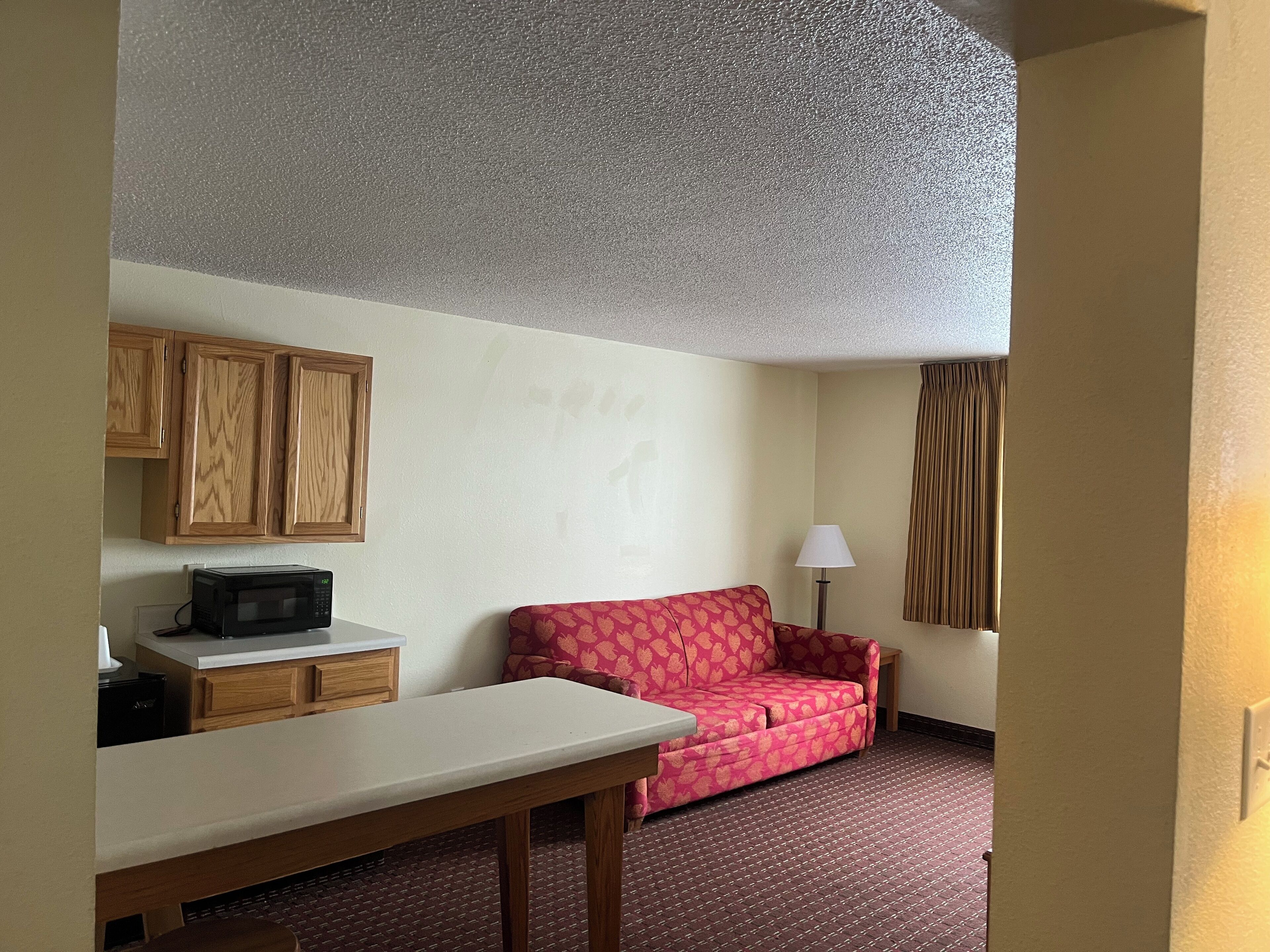 A hotel room with a kitchenette, a red patterned sofa, a lamp, and a ceiling with a textured finish.