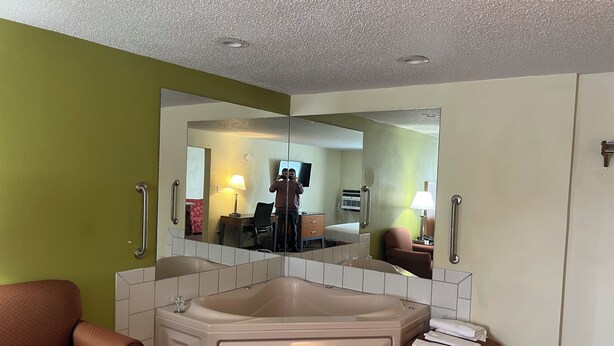 Une salle de bain d’hôtel avec un grand miroir, une baignoire, deux fauteuils et une petite table avec une lampe.