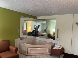 Un baño de hotel con un espejo grande, una bañera, dos sillones y una mesita de noche con lámpara.