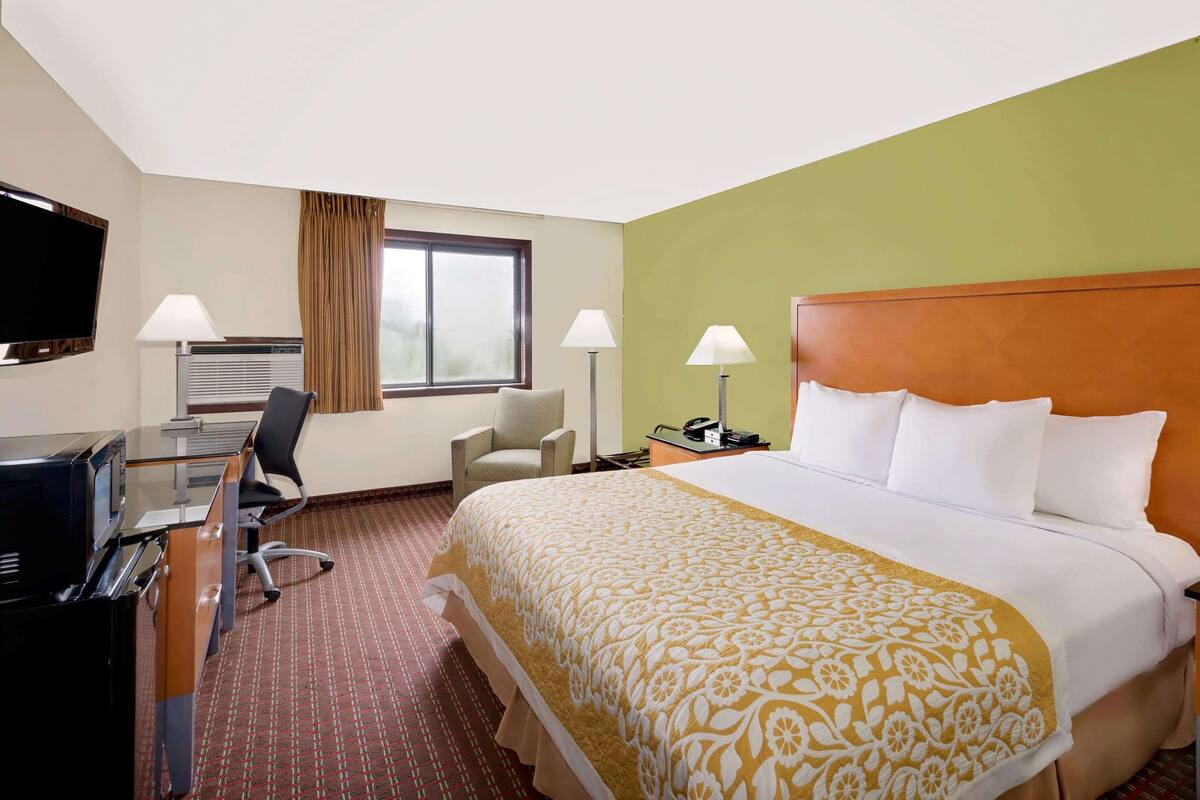A hotel room with a bed, two chairs, a desk with a computer, a TV, and a window with curtains.