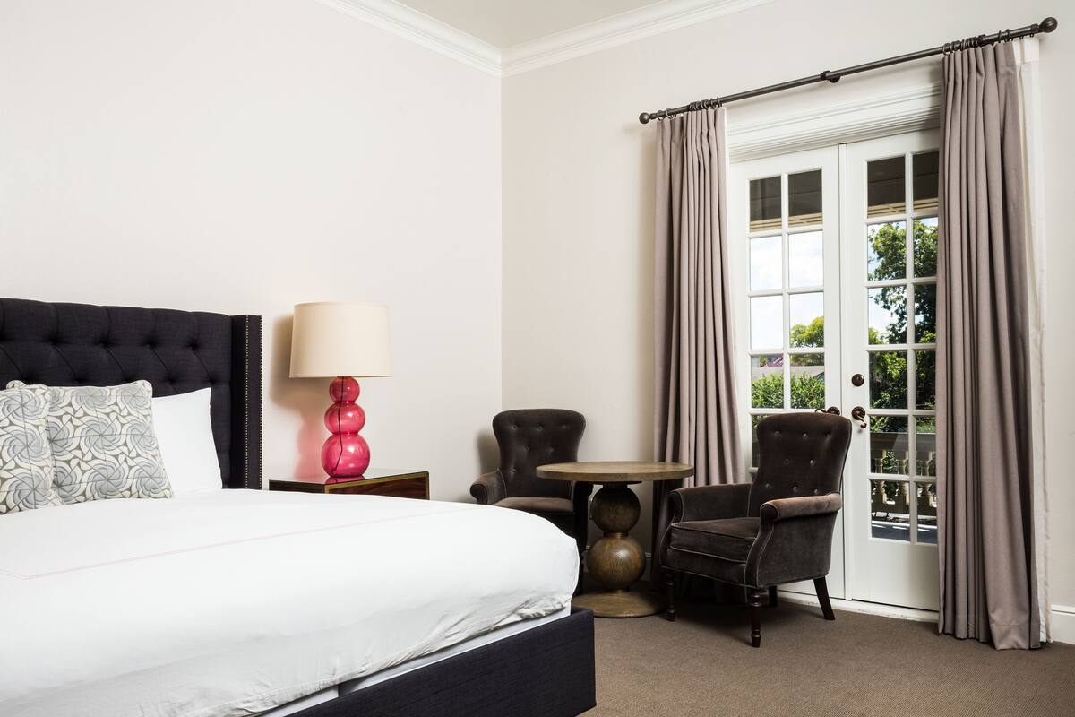 A hotel room with a large bed, a round table, two armchairs, a lamp, and a view of the outdoors through a window with curtains.