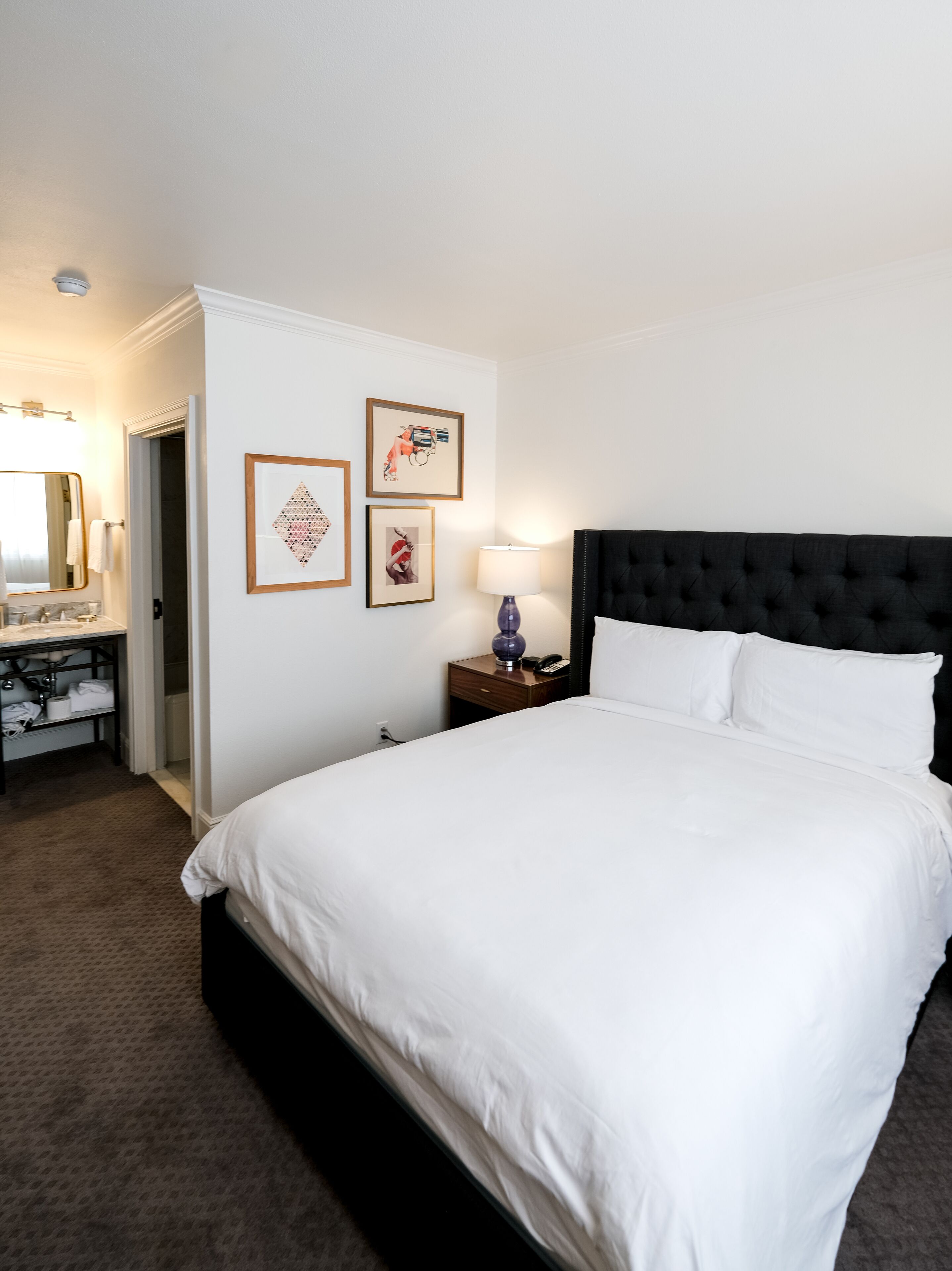 A hotel room with a large bed, a nightstand with a lamp, a dresser, and framed artwork on the wall.
