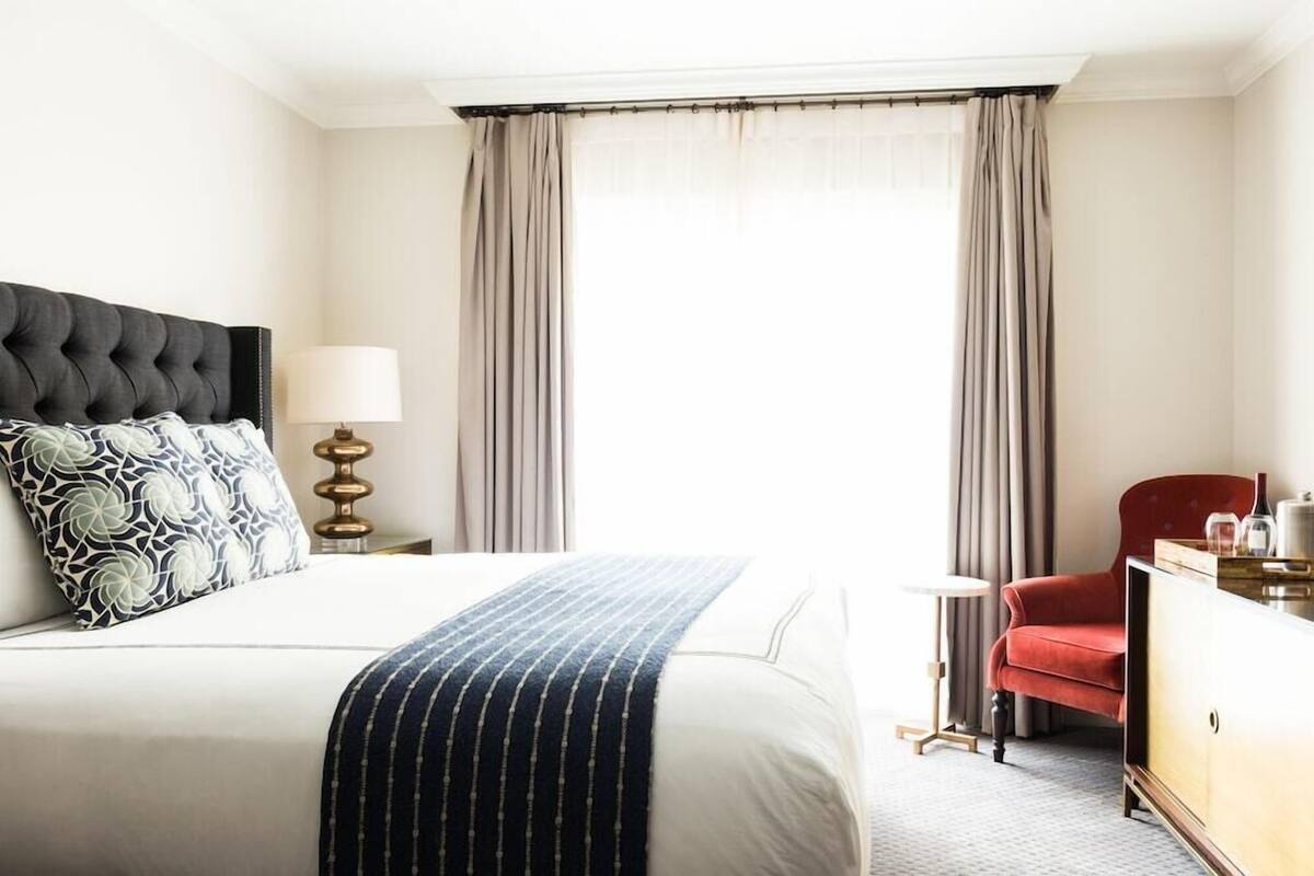 Habitación de hotel con una cama grande, un sillón rojo, una mesita de noche con una lámpara y vistas al exterior a través de cortinas traslúcidas.