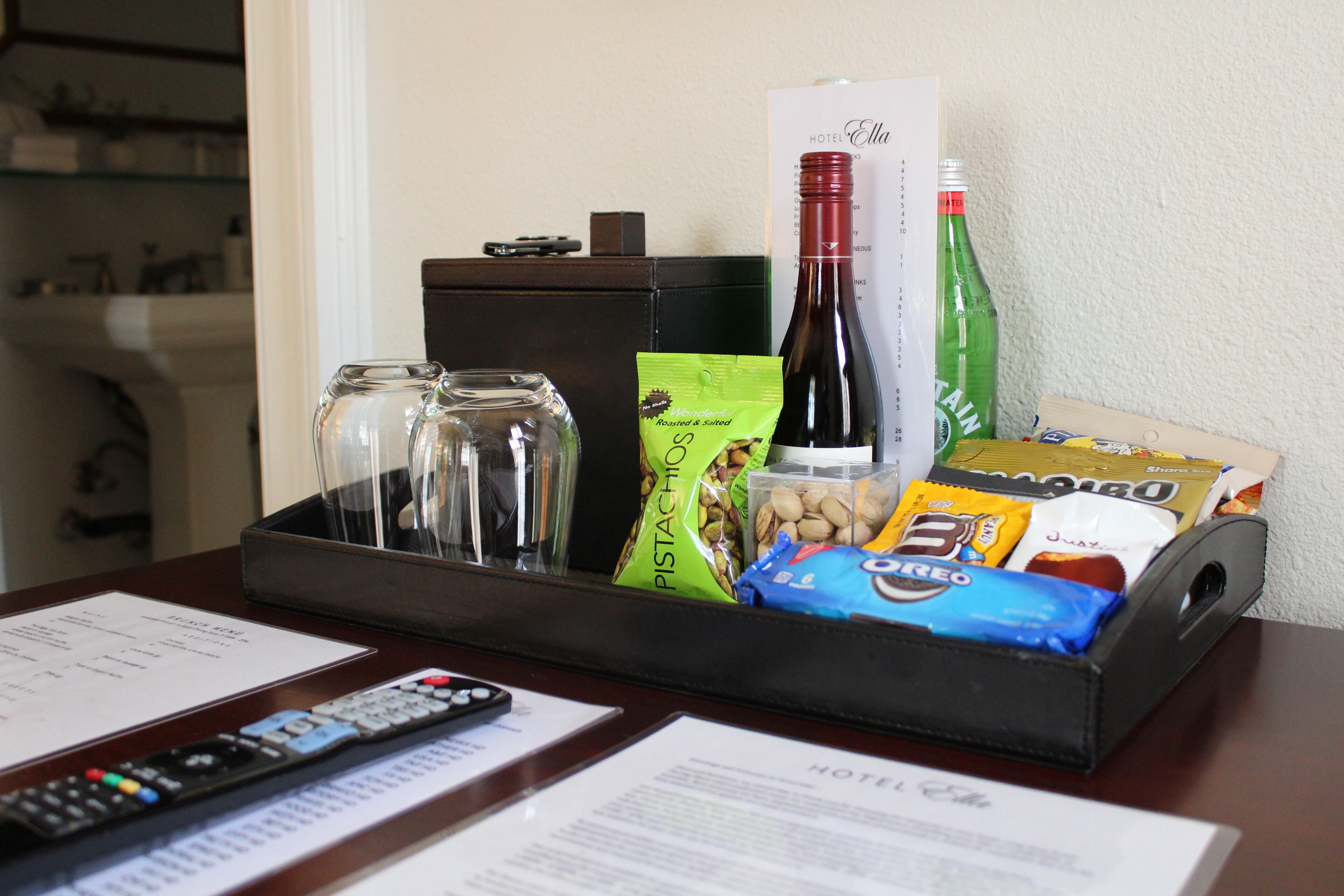 A hotel room's reception area with a tray containing a wine bottle, a green bottle, a packet of pistachios, a bag of Oreos, and a remote control.